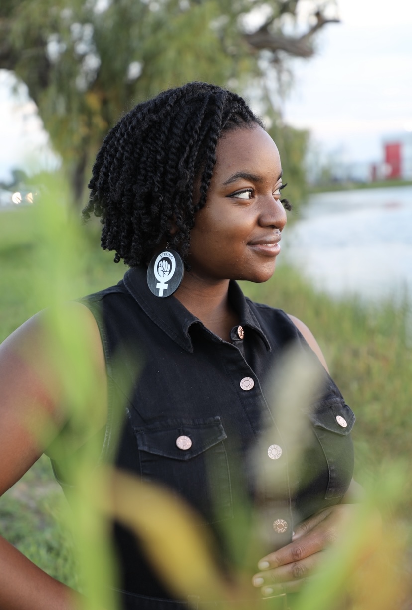 Young woman with natural curly hair outdoors, wearing a black shirt with pink buttons, near a body of water.