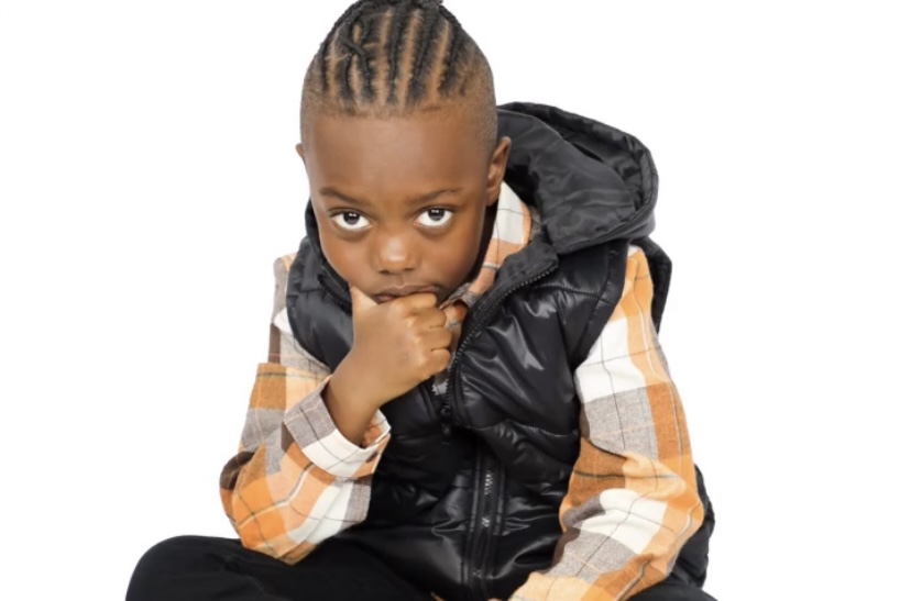 Child with braided hair wearing a black vest and plaid shirt, sitting with hand near mouth, looking at camera.