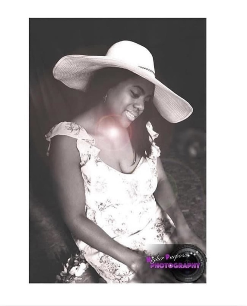 Young girl wearing a large sun hat and floral dress, smiling with eyes closed, sitting outdoors. Black and white photo.