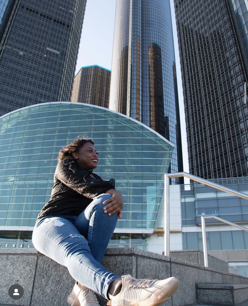 Woman sitting on steps in front of modern glass skyscrapers, smiling and looking up, wearing a black jacket and light jeans.