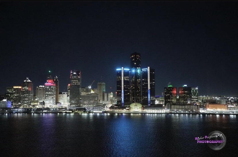 Nighttime city skyline with illuminated buildings reflected on water, dark sky, and bright lights on skyscrapers.