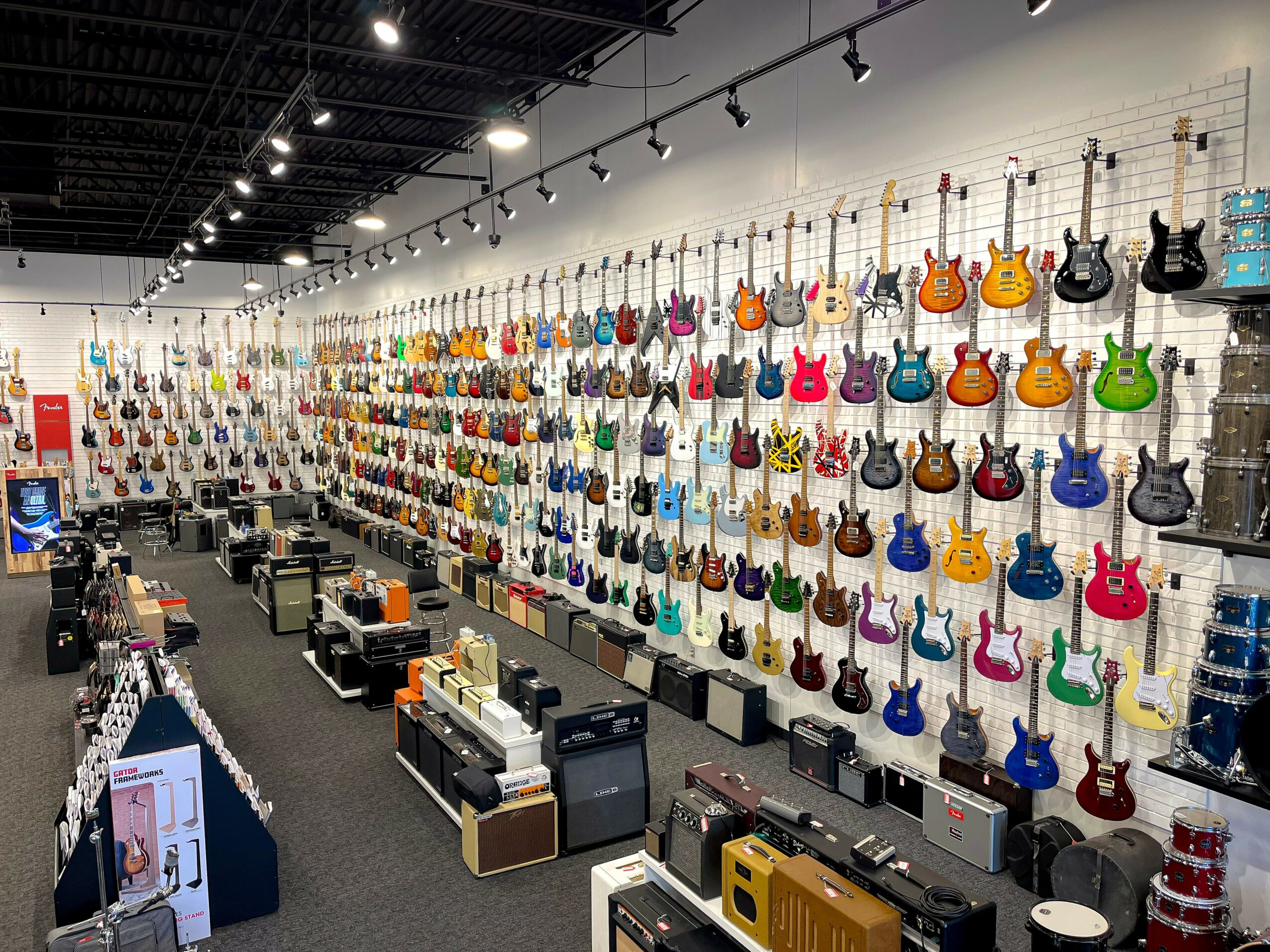 Wall filled with colorful electric guitars displayed on hooks in a music store, with counters and equipment below.