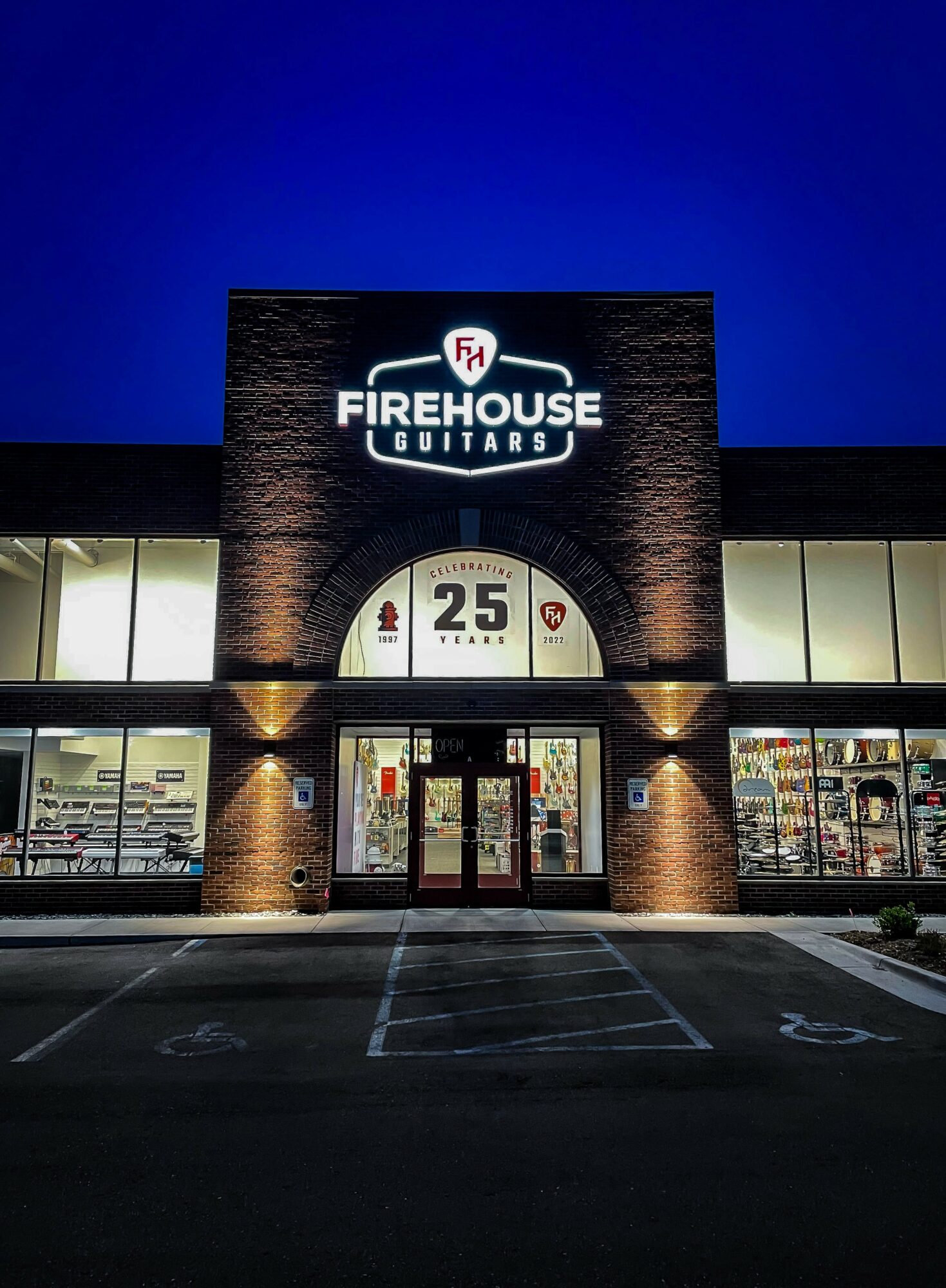 Storefront with illuminated sign reading 'Firehouse Guitars' against a dark blue sky, brick facade, and large glass windows.