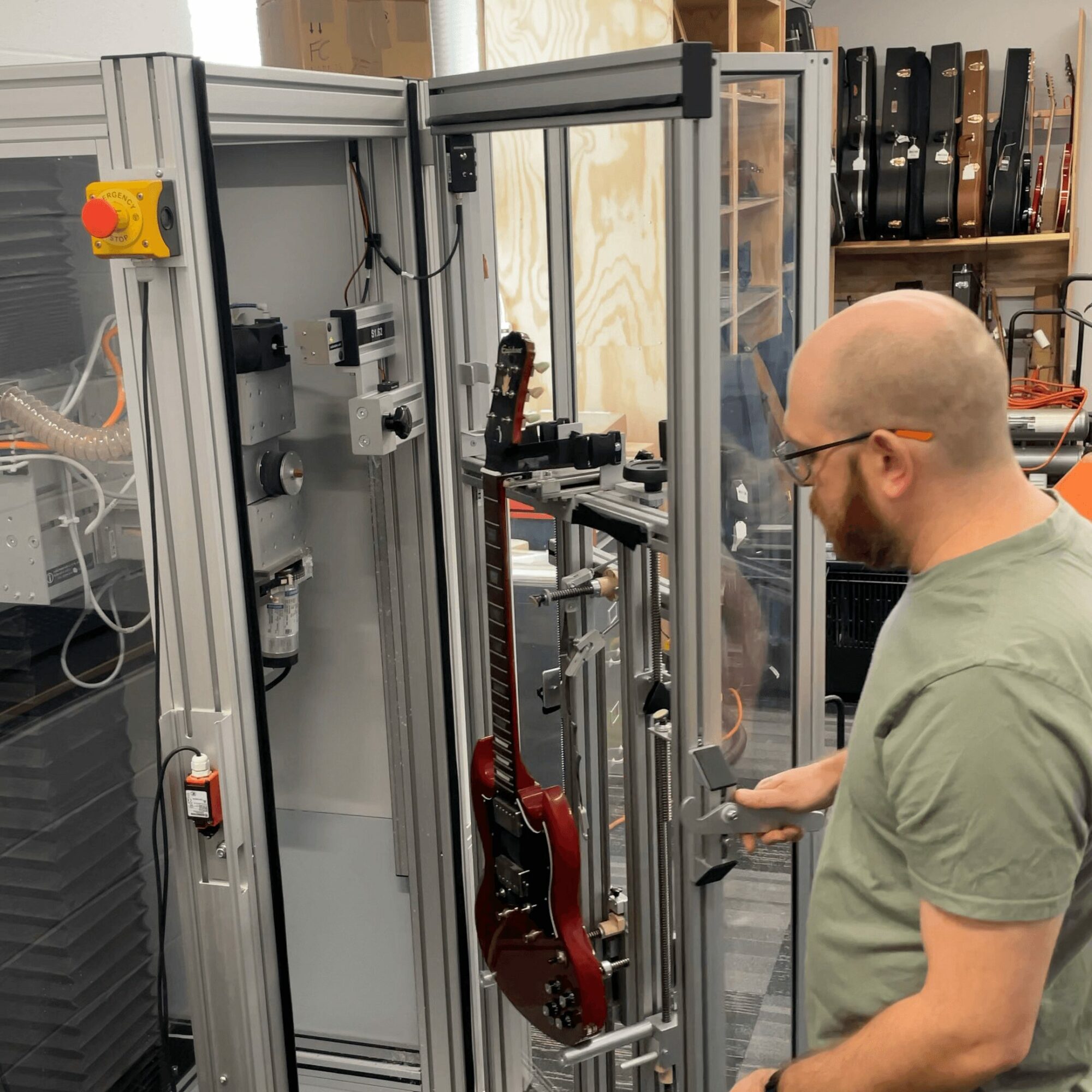 Man with glasses and a beard working on a machine with a red guitar inside a laboratory or workshop.