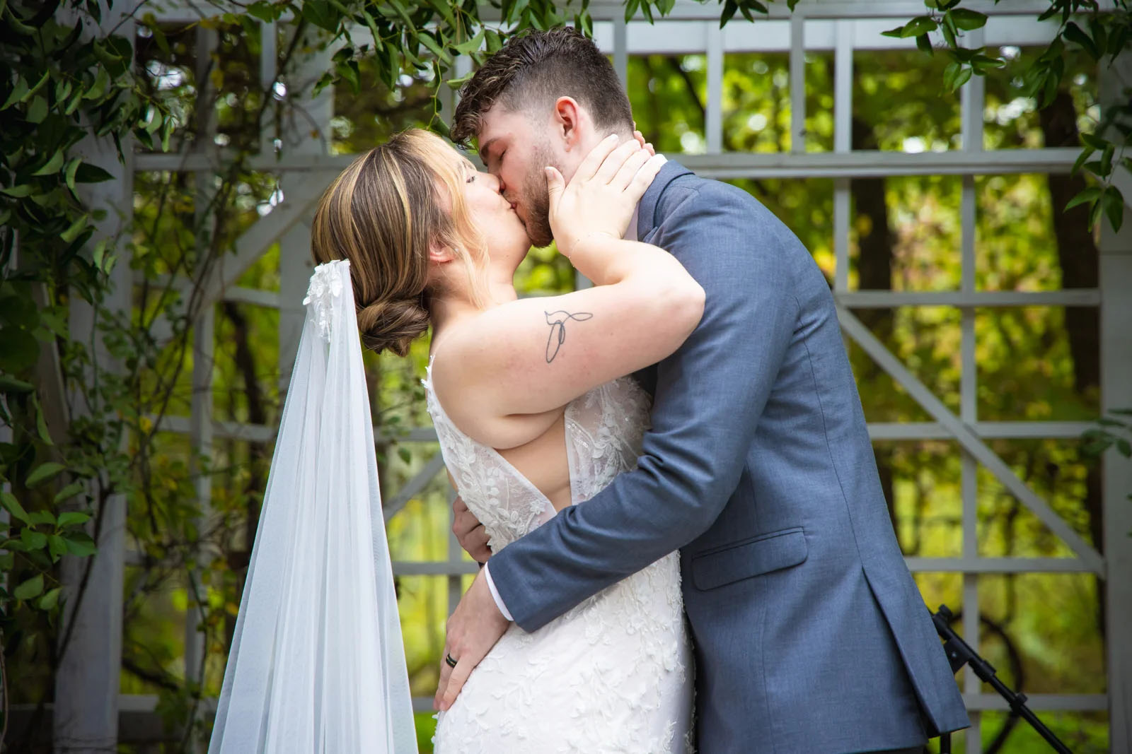 Couple kissing outdoors, woman in wedding dress and veil, man in blue suit, greenery background.