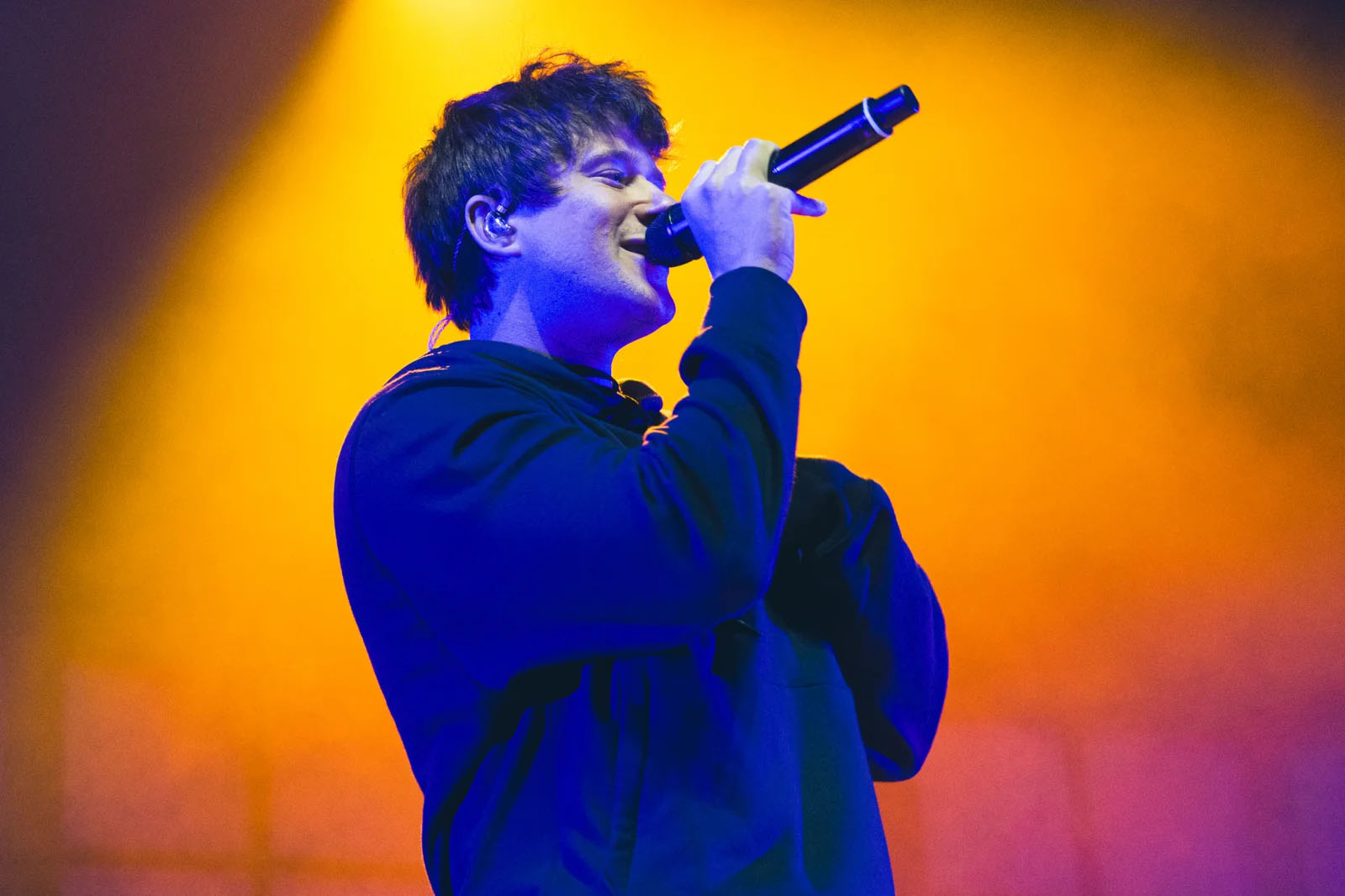 Person singing into a microphone with yellow and orange background, wearing a dark hoodie.