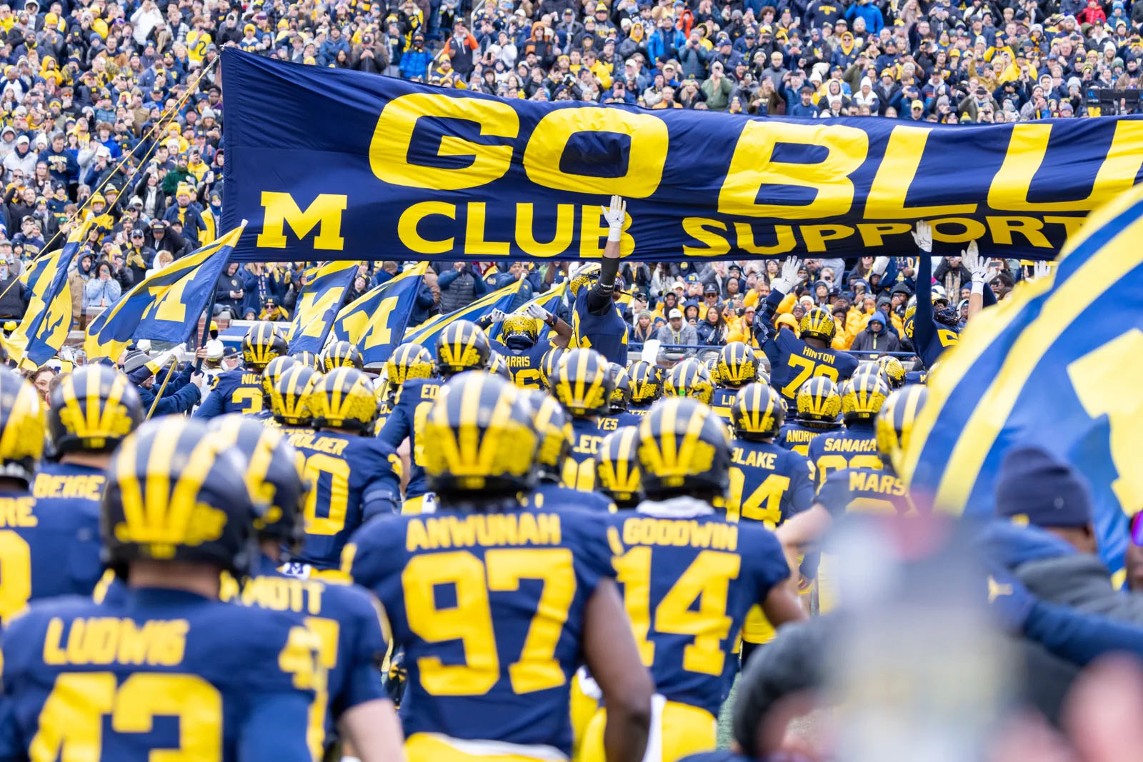Football team in blue and yellow uniforms with a large blue banner reading 'GO BLUE' and 'M CLUB SUPPORT' in a stadium.