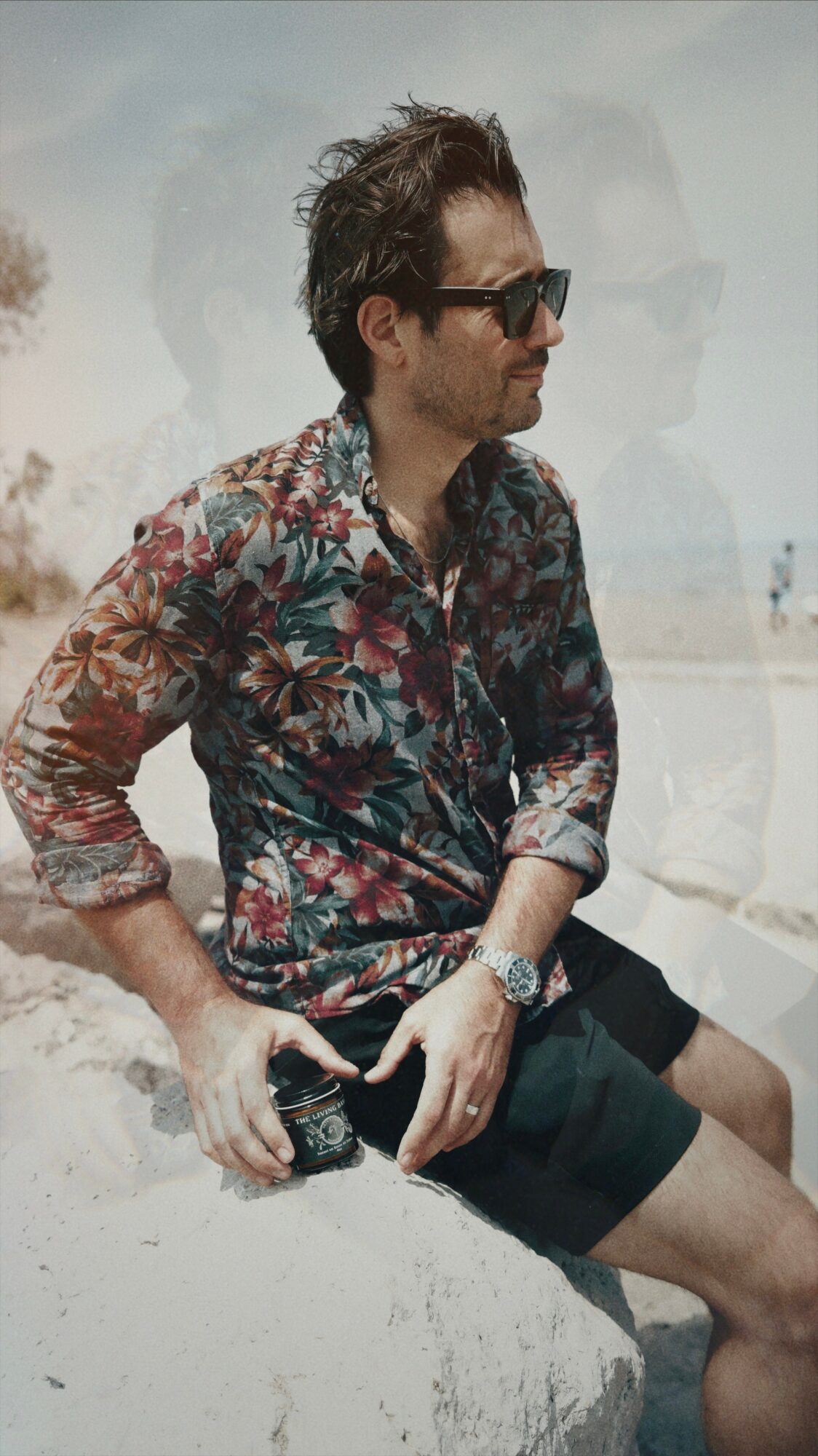 Man sitting outdoors wearing sunglasses, a floral shirt, and shorts, holding a camera, with a beach background.