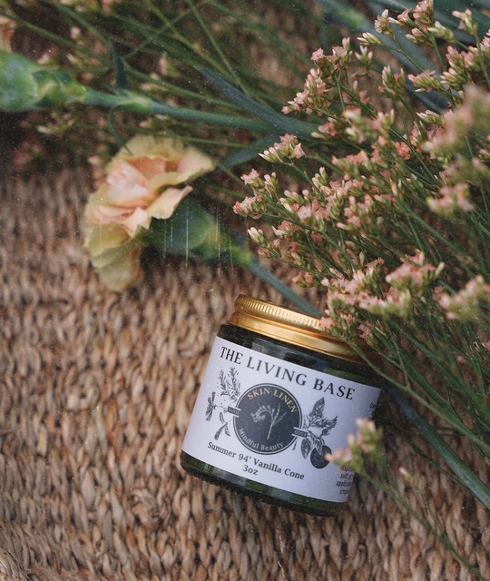 A small jar labeled 'The Living Base' placed on a woven surface surrounded by pink and green flowers.