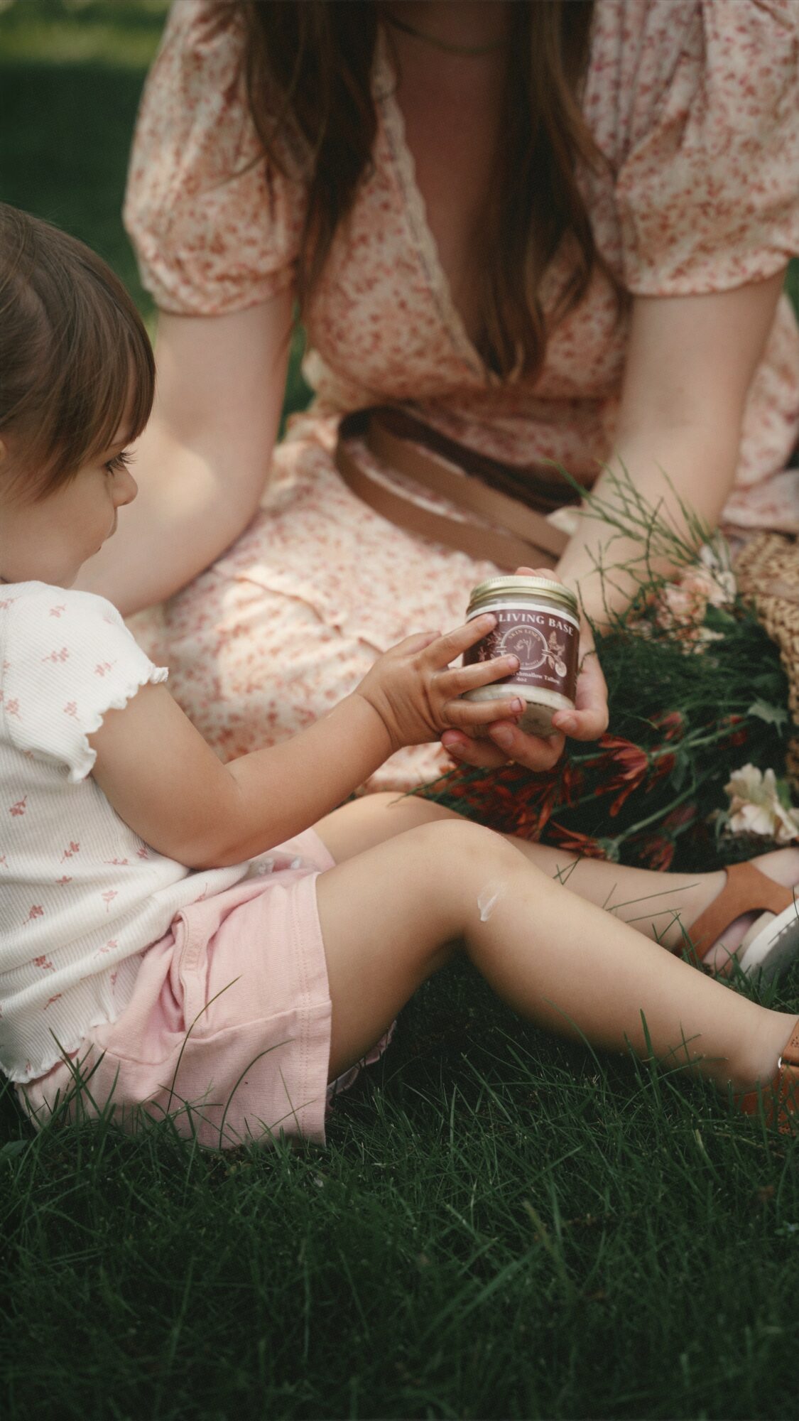 Child and woman sitting on grass, child holding a jar, woman wearing a pink dress with brown belt, greenery in background.