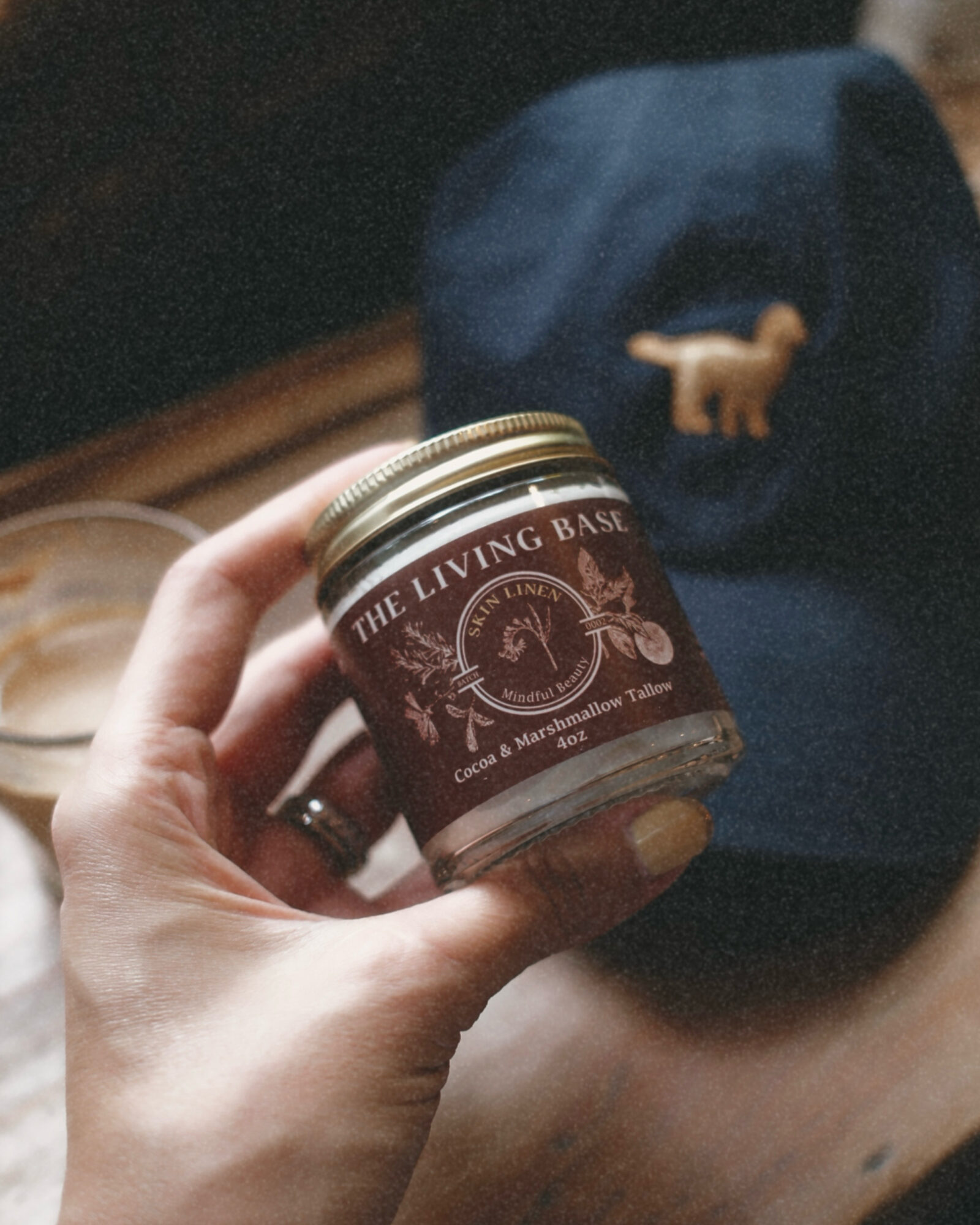 Hand holding a small jar labeled 'The Living Bast' with a dark background and a blue cap with a dog pin.