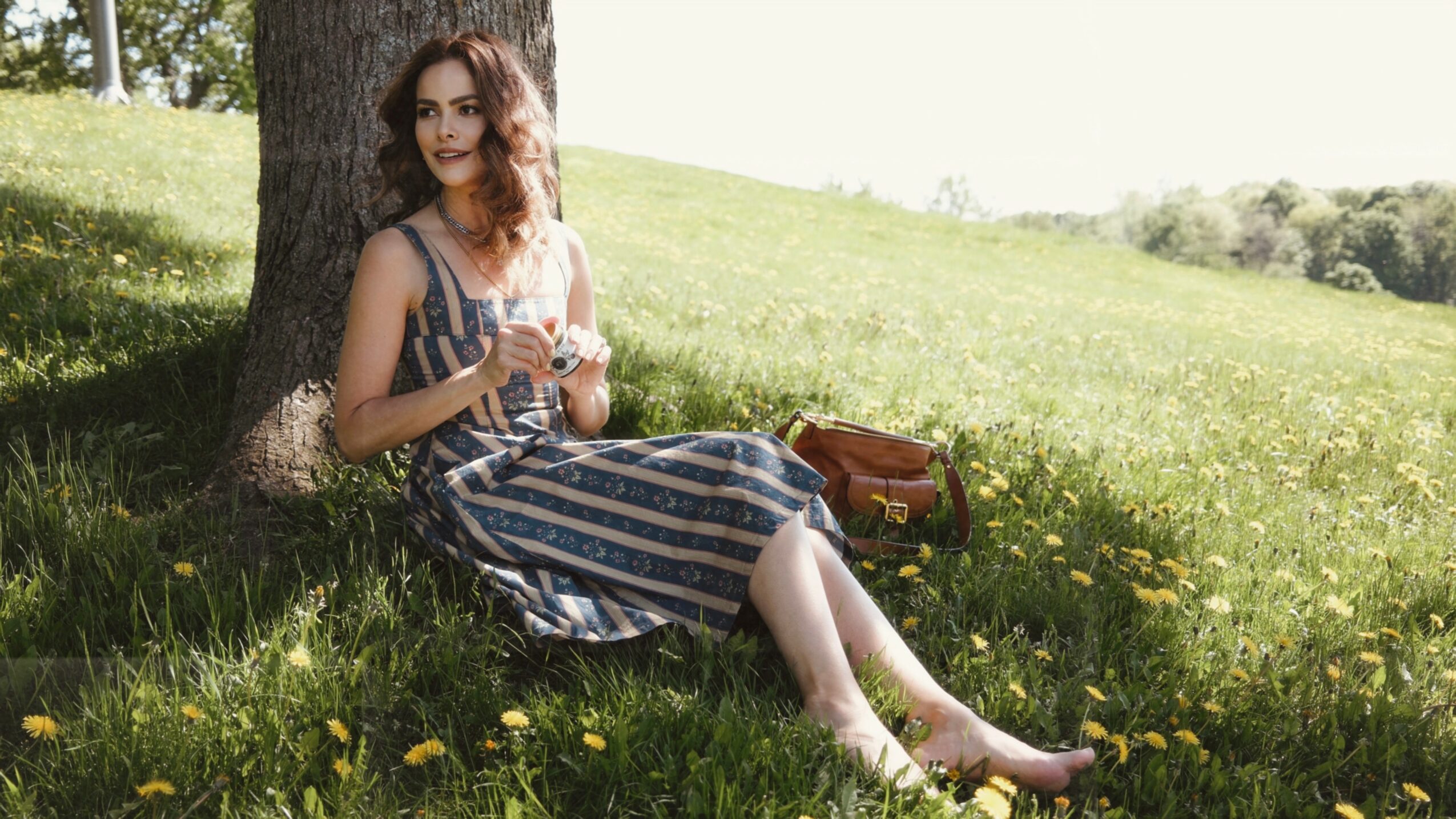 Woman sitting under a tree in a grassy field, holding a cup, wearing a striped dress, with a bag nearby.