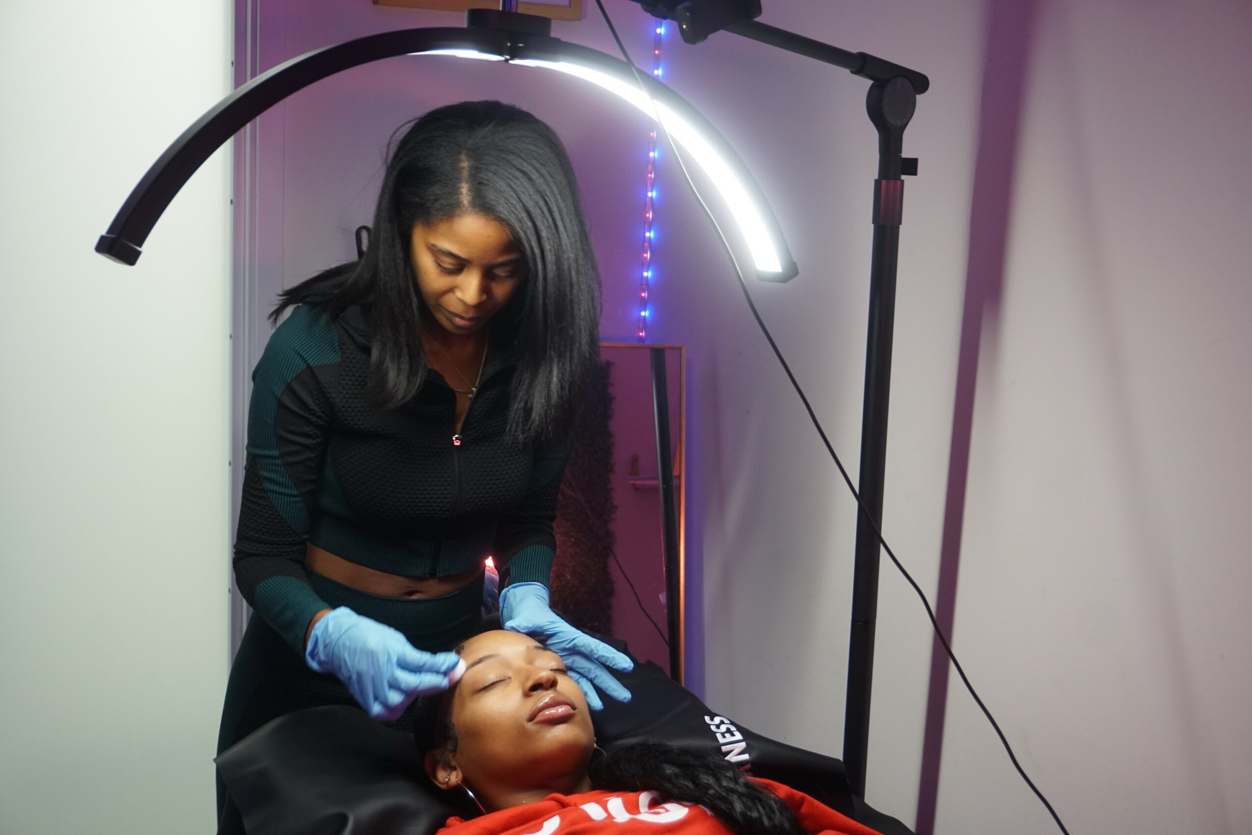 A woman in gloves performs a facial treatment on a woman lying down, under a bright light in a clinical setting.