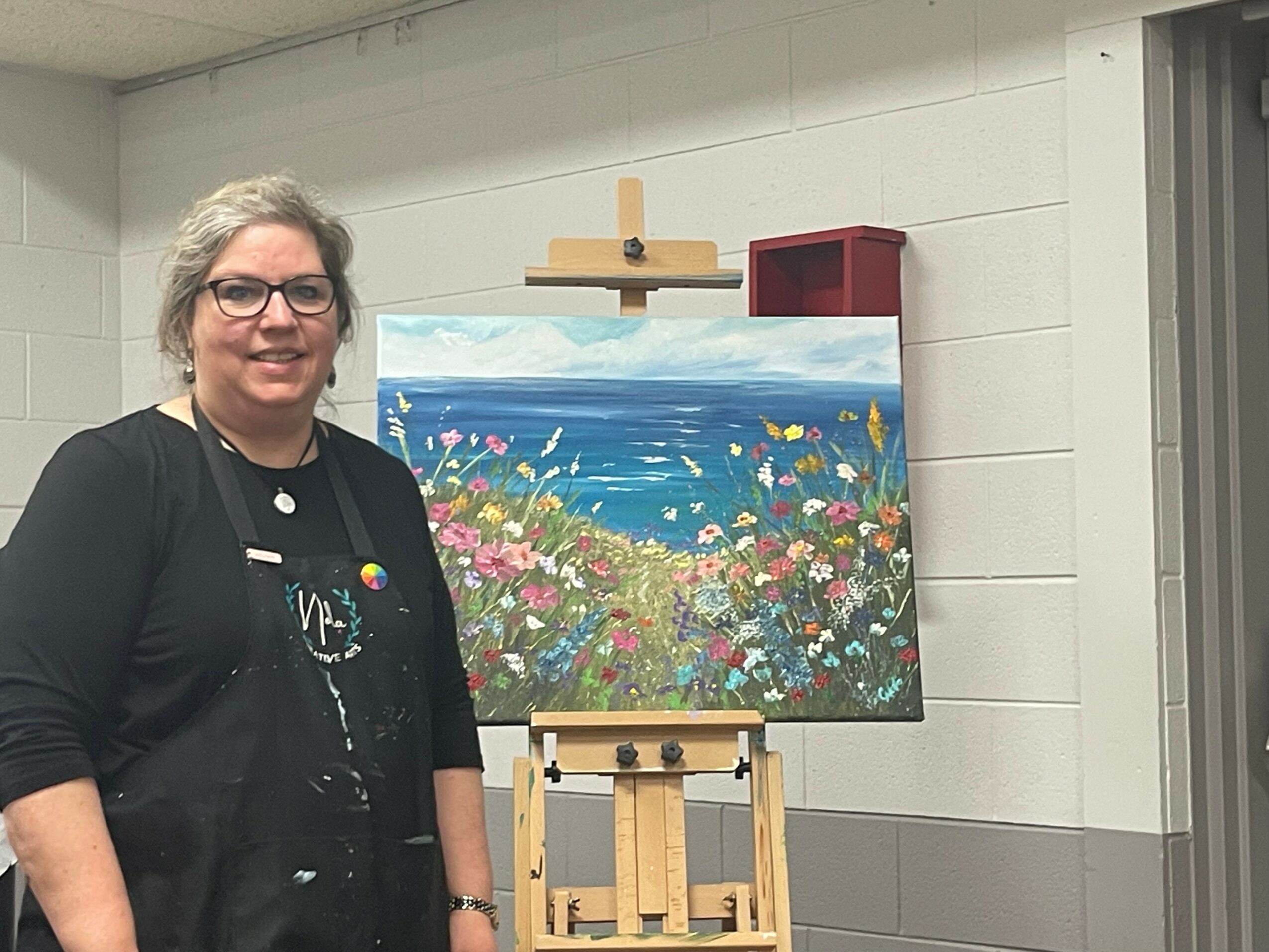 Woman with glasses and black clothing standing next to a colorful landscape painting on an easel in a room.