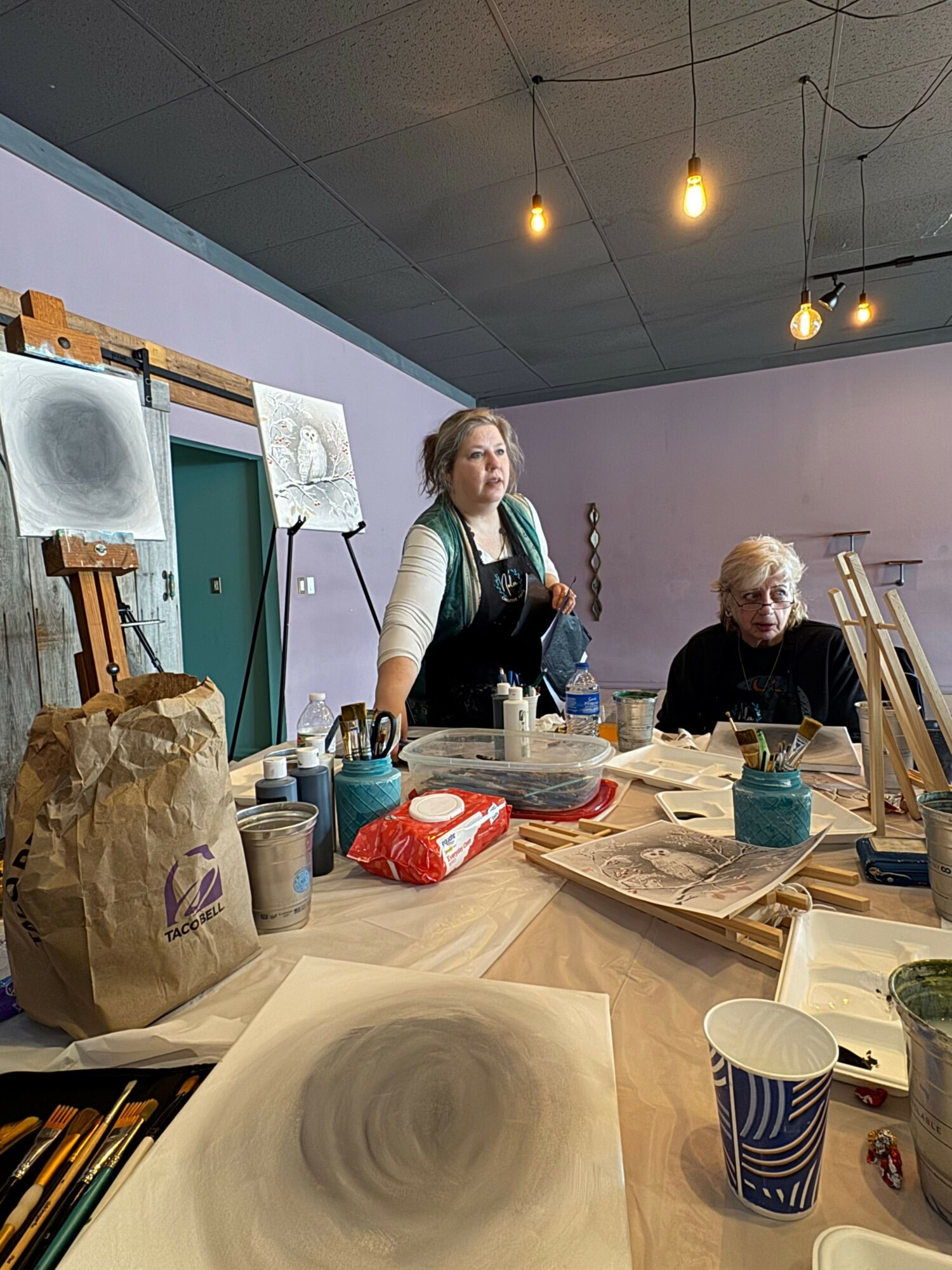 Two women sit at a table with food and drinks, in a room with a high ceiling and hanging lights.