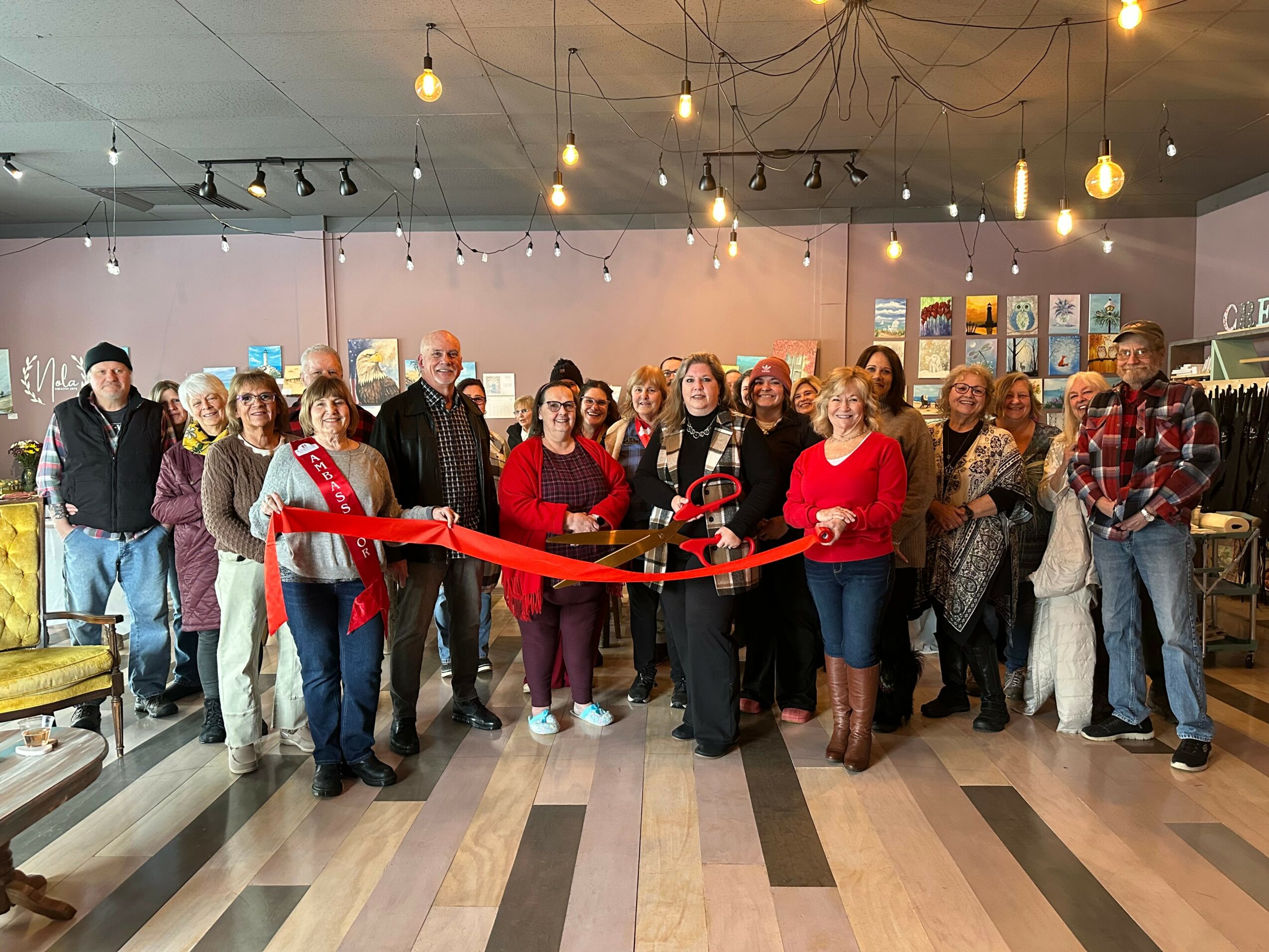 Group of people gathered indoors for a ribbon-cutting event, with a woman holding scissors and a red ribbon, in a decorated space.