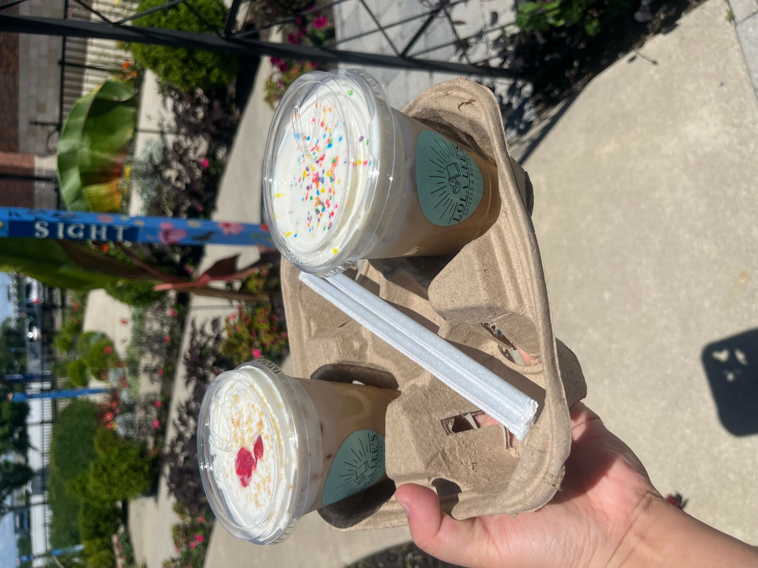 Hand holding a tray with two bubble tea drinks, straws, and a napkin, outdoors on a sidewalk.