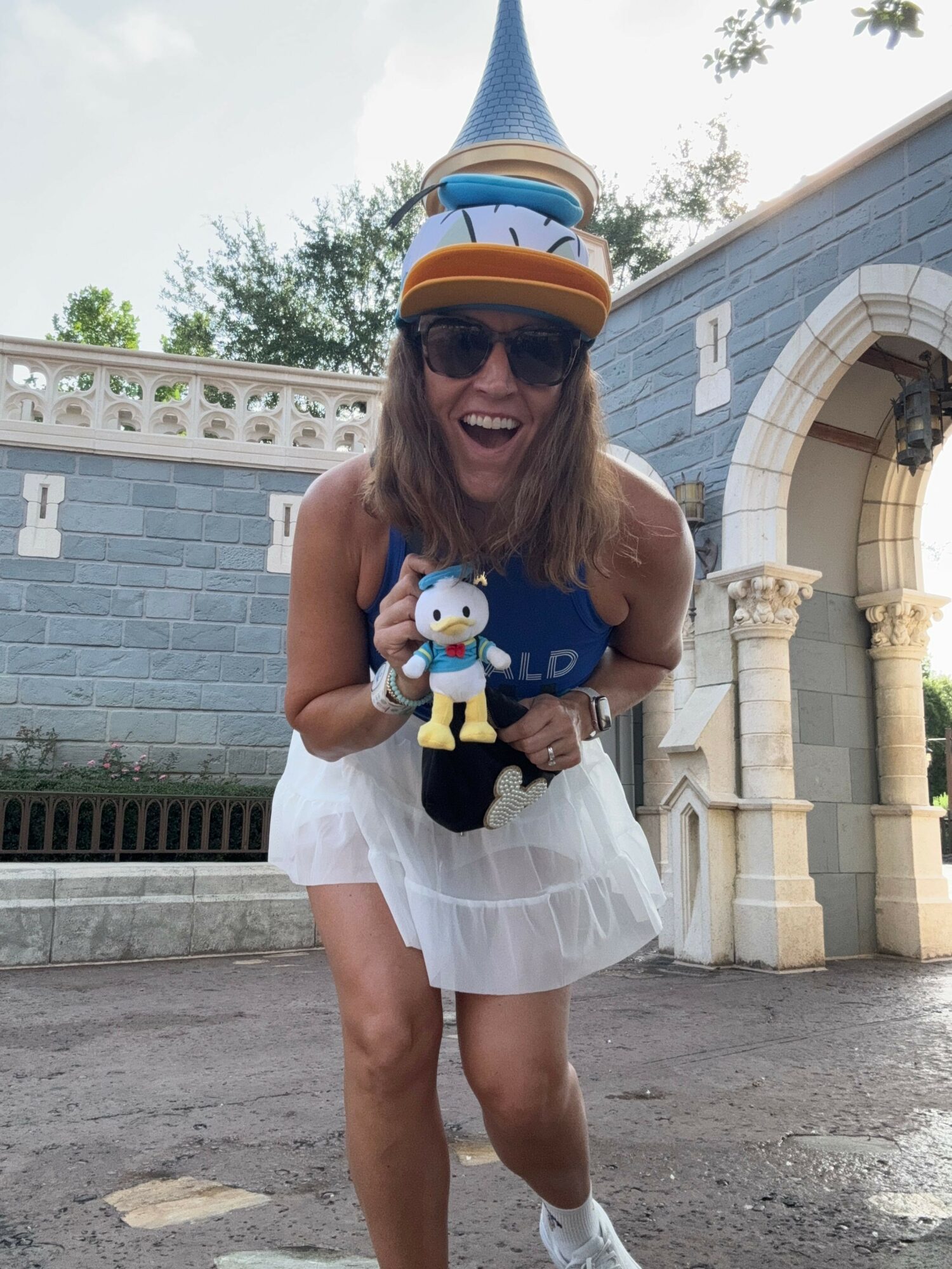 Person wearing sunglasses and a hat, holding a snowman figure, standing outdoors near an arched entrance with trees in the background.