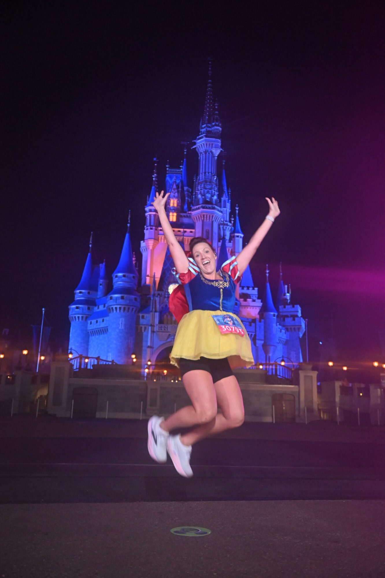 Person jumping in front of a brightly lit castle at night, arms raised, smiling, wearing colorful clothing.