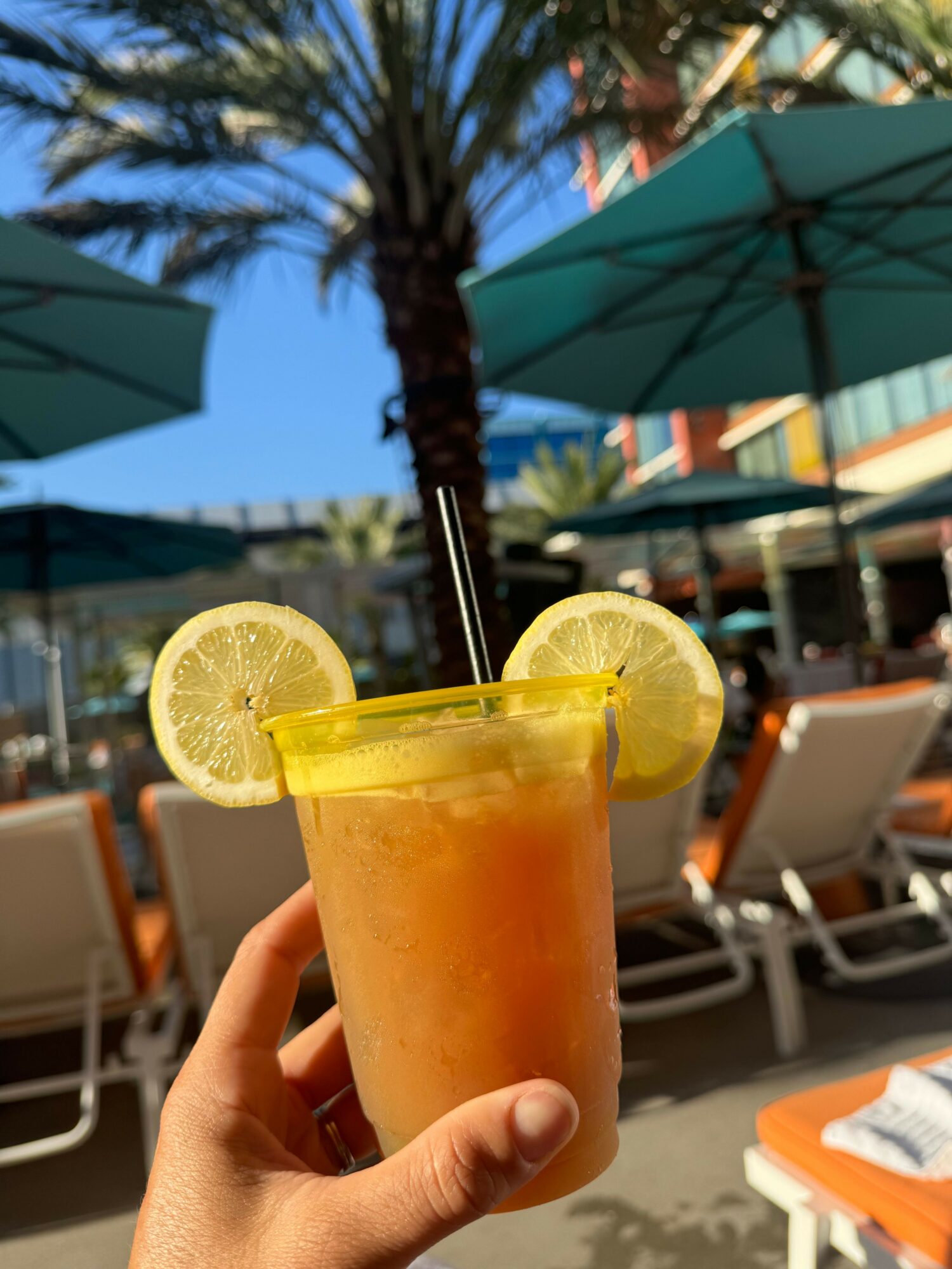 Person holding a glass of iced drink with lemon slices at poolside area with umbrellas and lounge chairs.