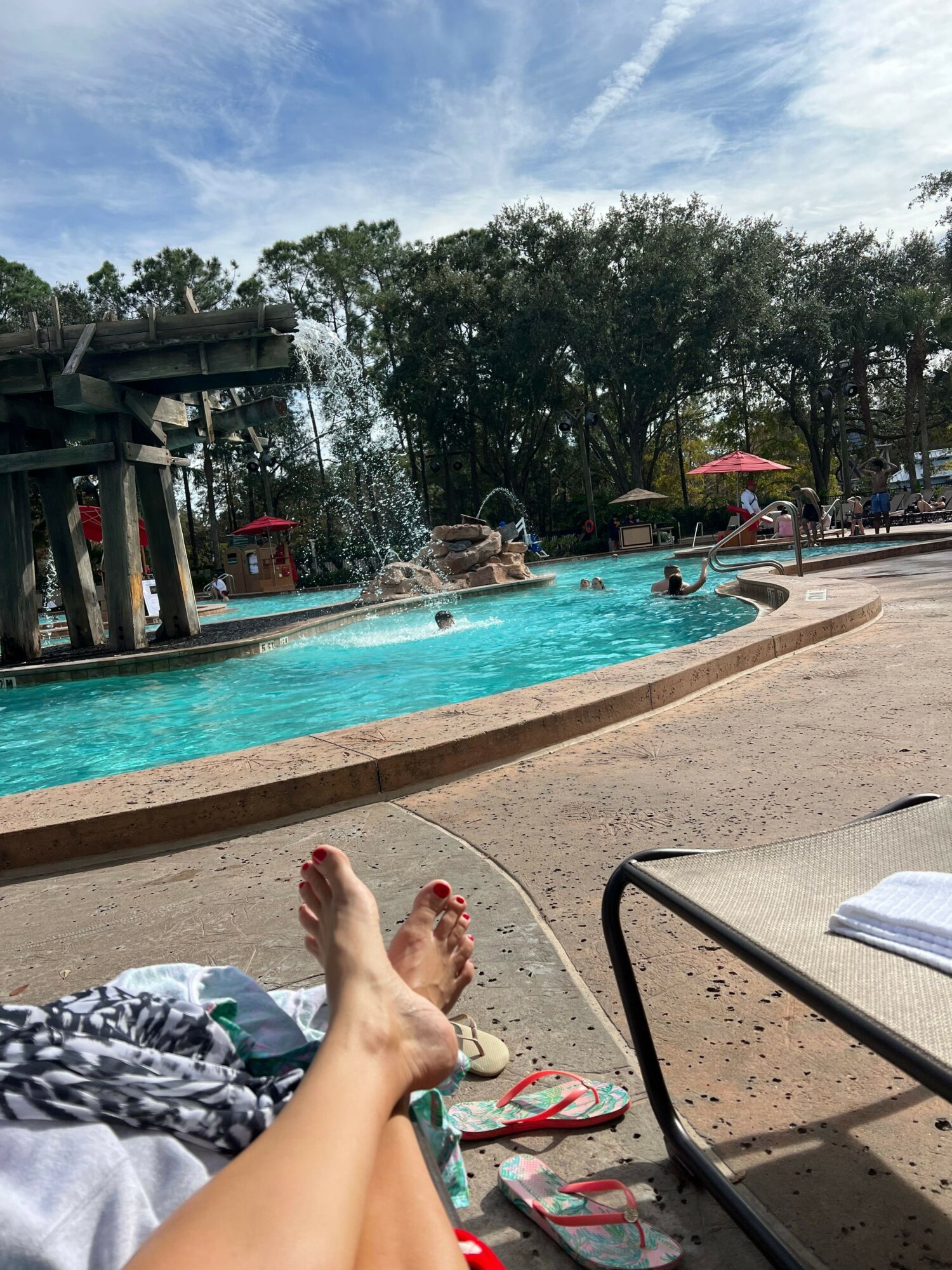 Person's legs and feet are visible near a swimming pool with lounge chairs and trees in the background.