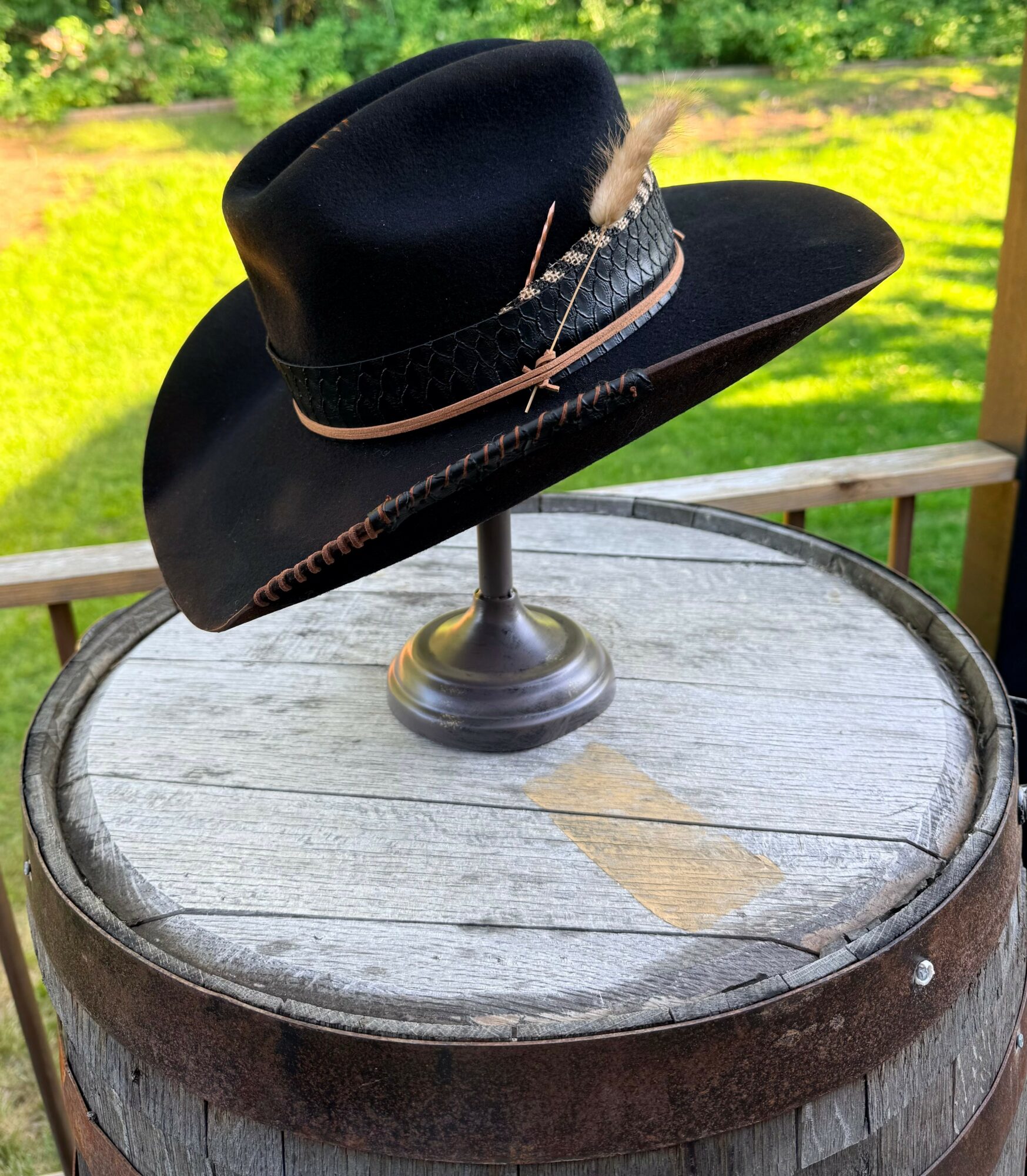 Black hat with feather and band on a stand, placed on a wooden barrel outdoors, green grass in background.