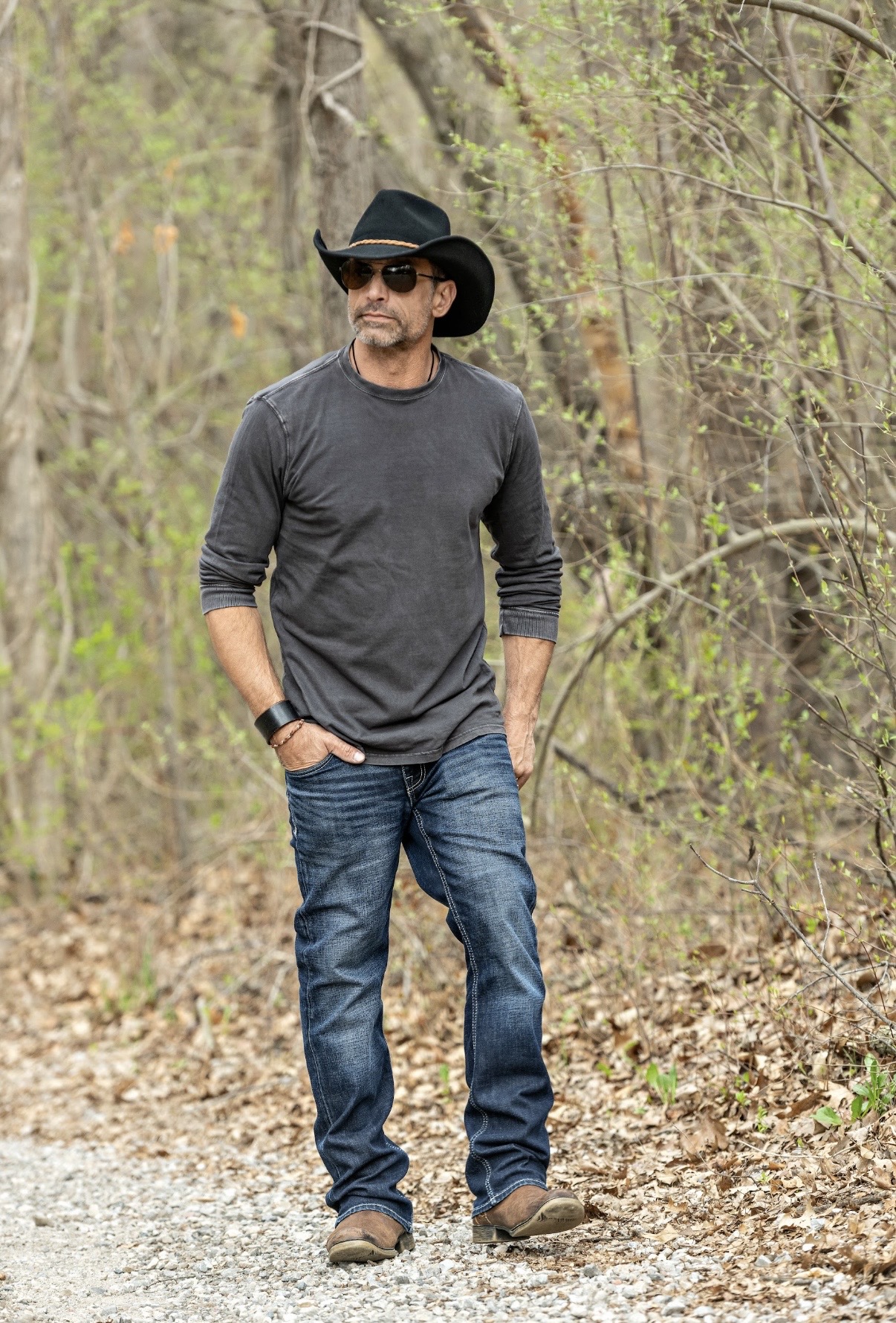 Man wearing sunglasses, a black hat, dark gray long-sleeve shirt, jeans, standing outdoors in a wooded area.