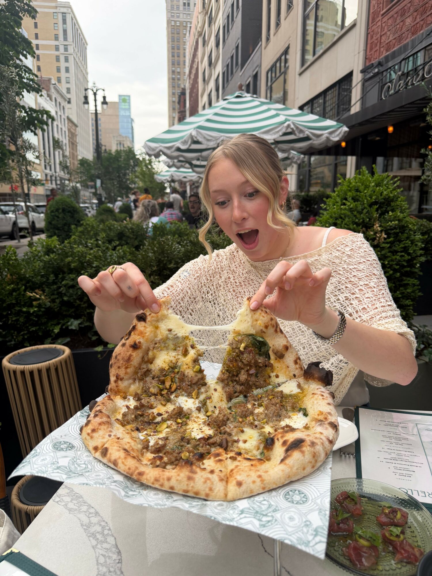 Woman with blonde hair and striped hat giving thumbs up next to pizza on table outdoors.