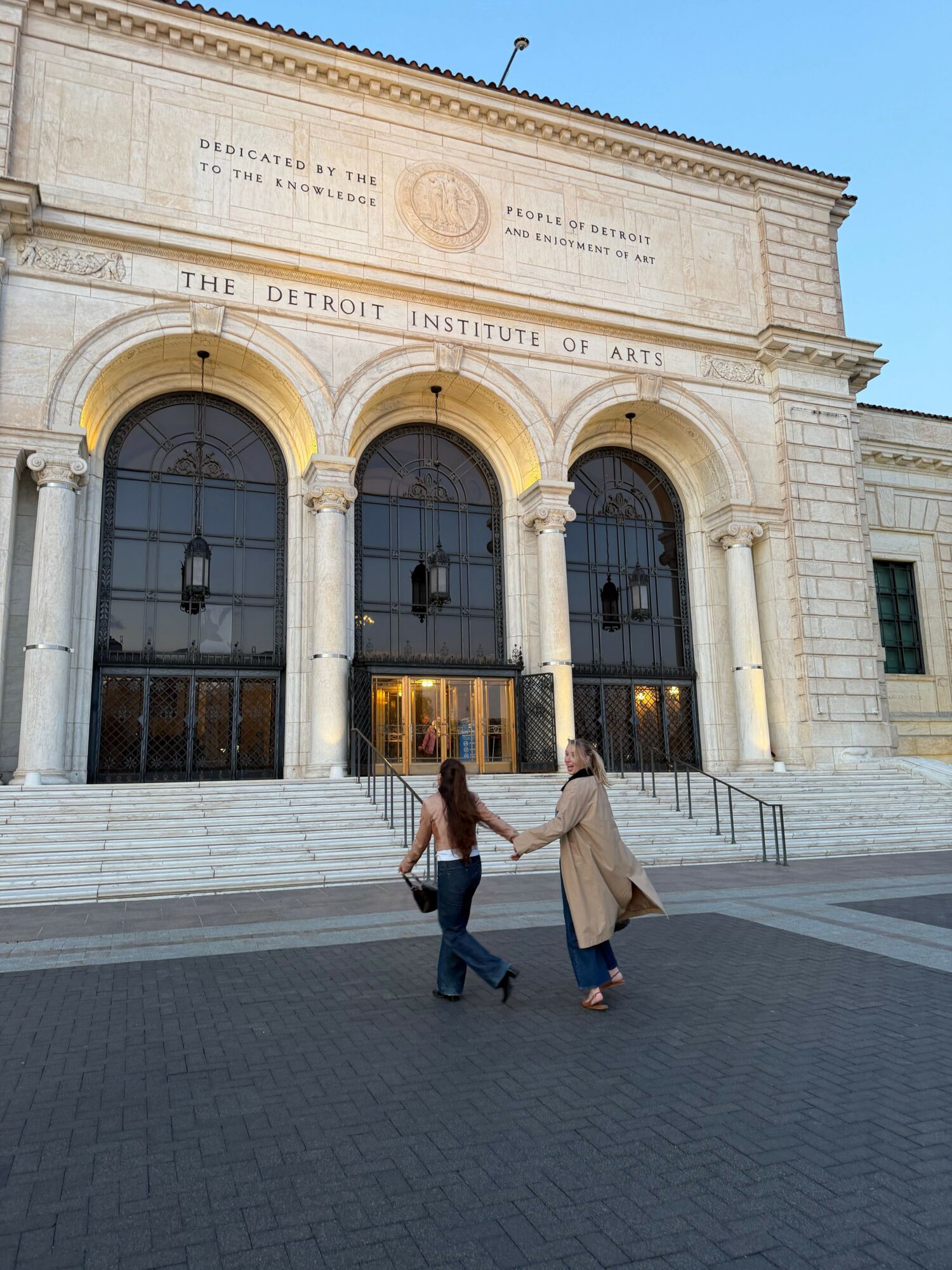 Two people walk past the entrance of a historic building with arched windows and stone architecture.