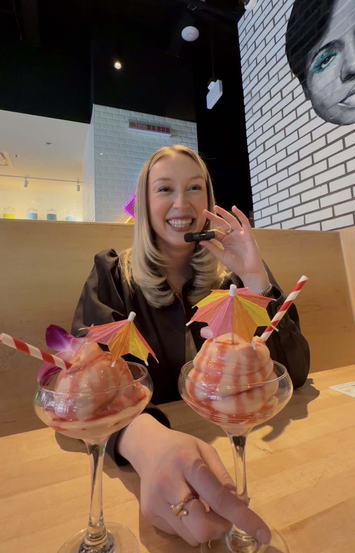 Smiling woman with blonde hair enjoying drinks with umbrellas and straws at a restaurant, pointing at the camera.