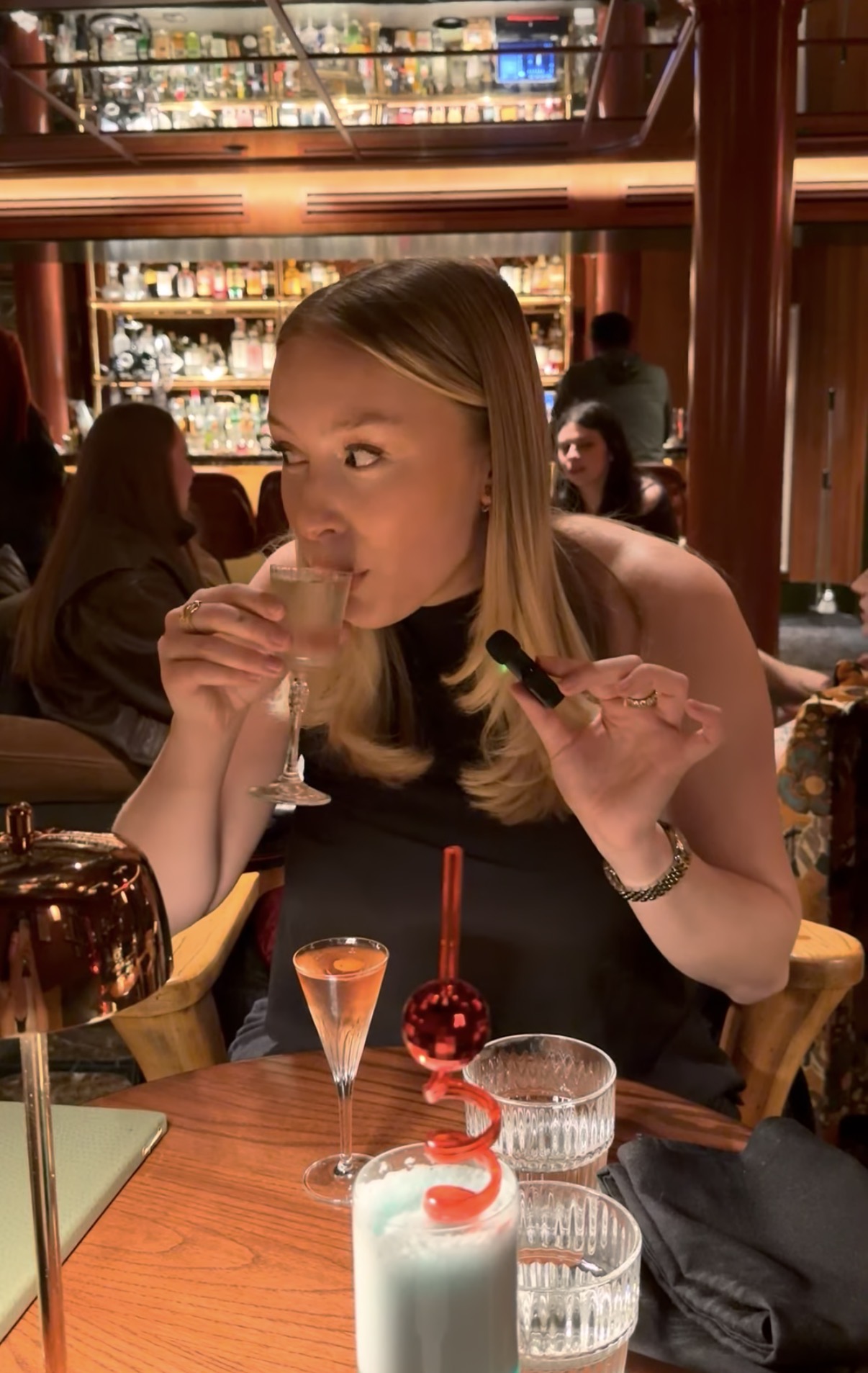 Woman sitting at a table in a restaurant, holding a vape device, with drinks and a birthday cake in front of her.