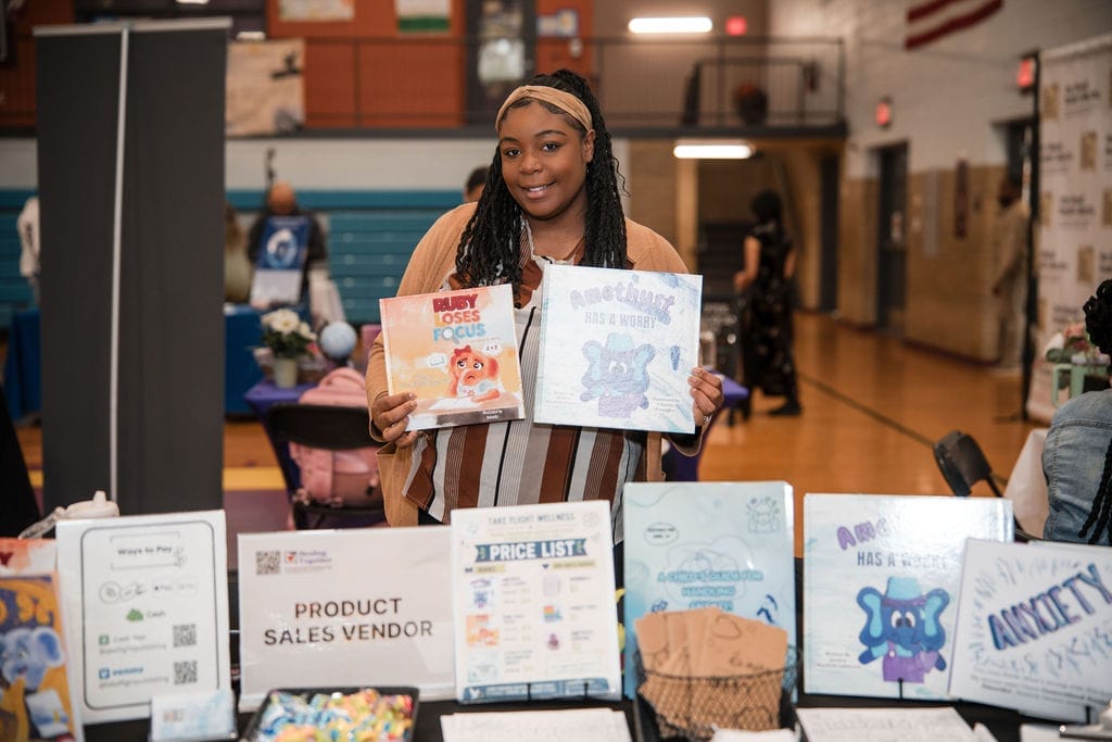 Young girl holding two books in a school gymnasium with tables and posters around her.