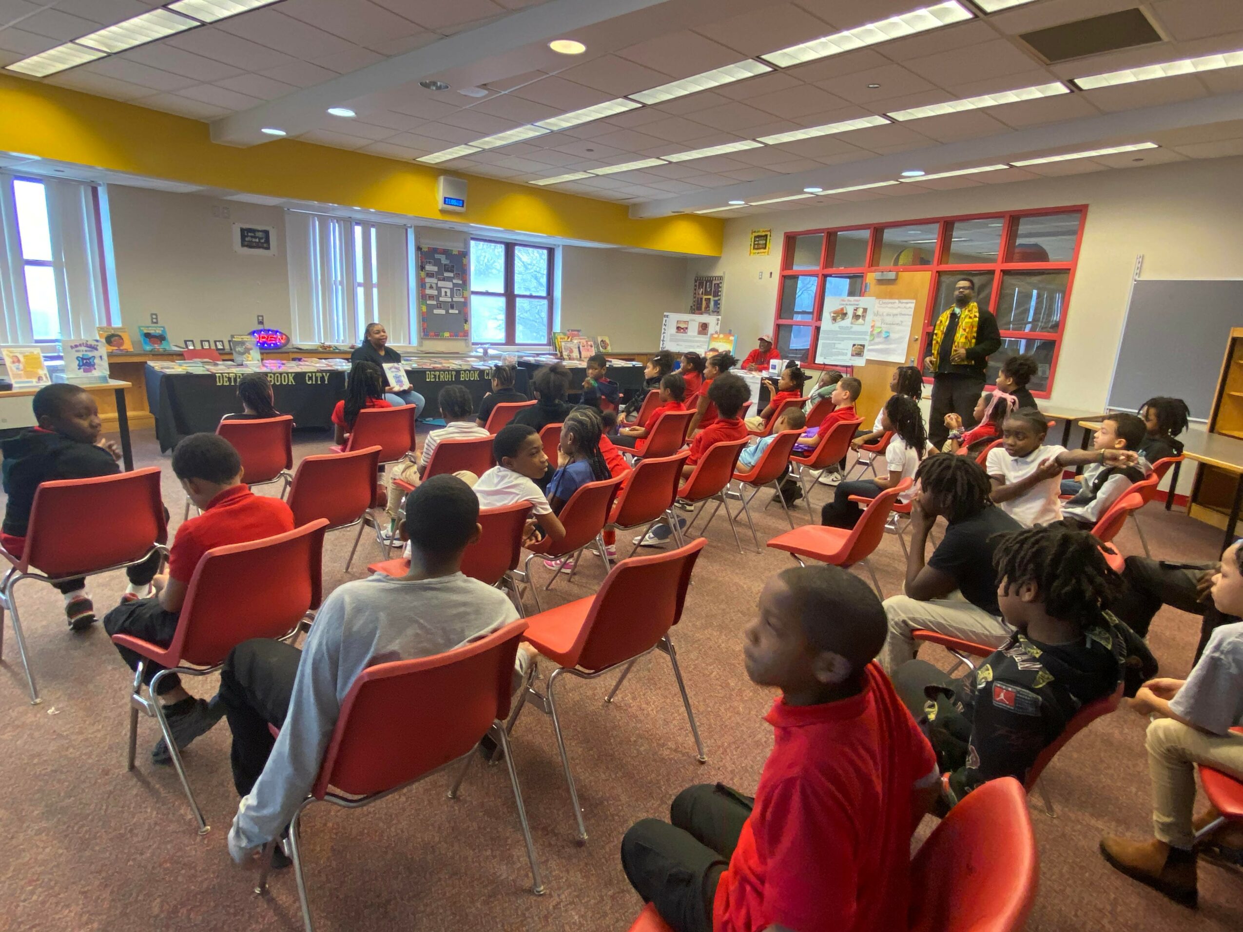 Children seated in a classroom, facing forward, with a teacher at the front near a whiteboard, windows on the left, and colorful walls.