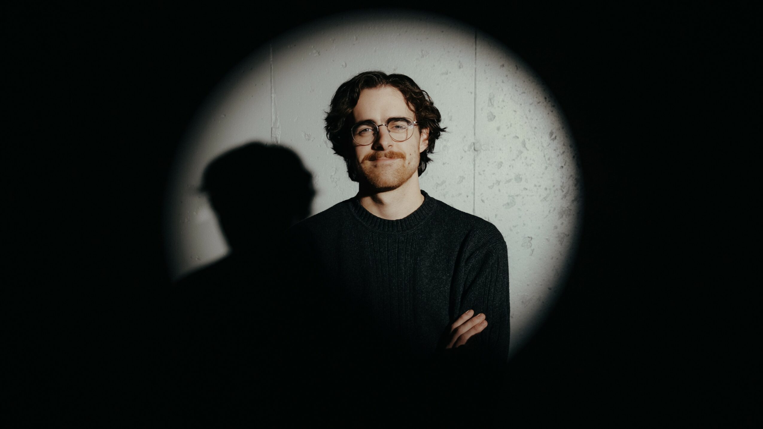 Person with glasses and curly hair smiling, standing against a light background, shadow cast on the wall behind.