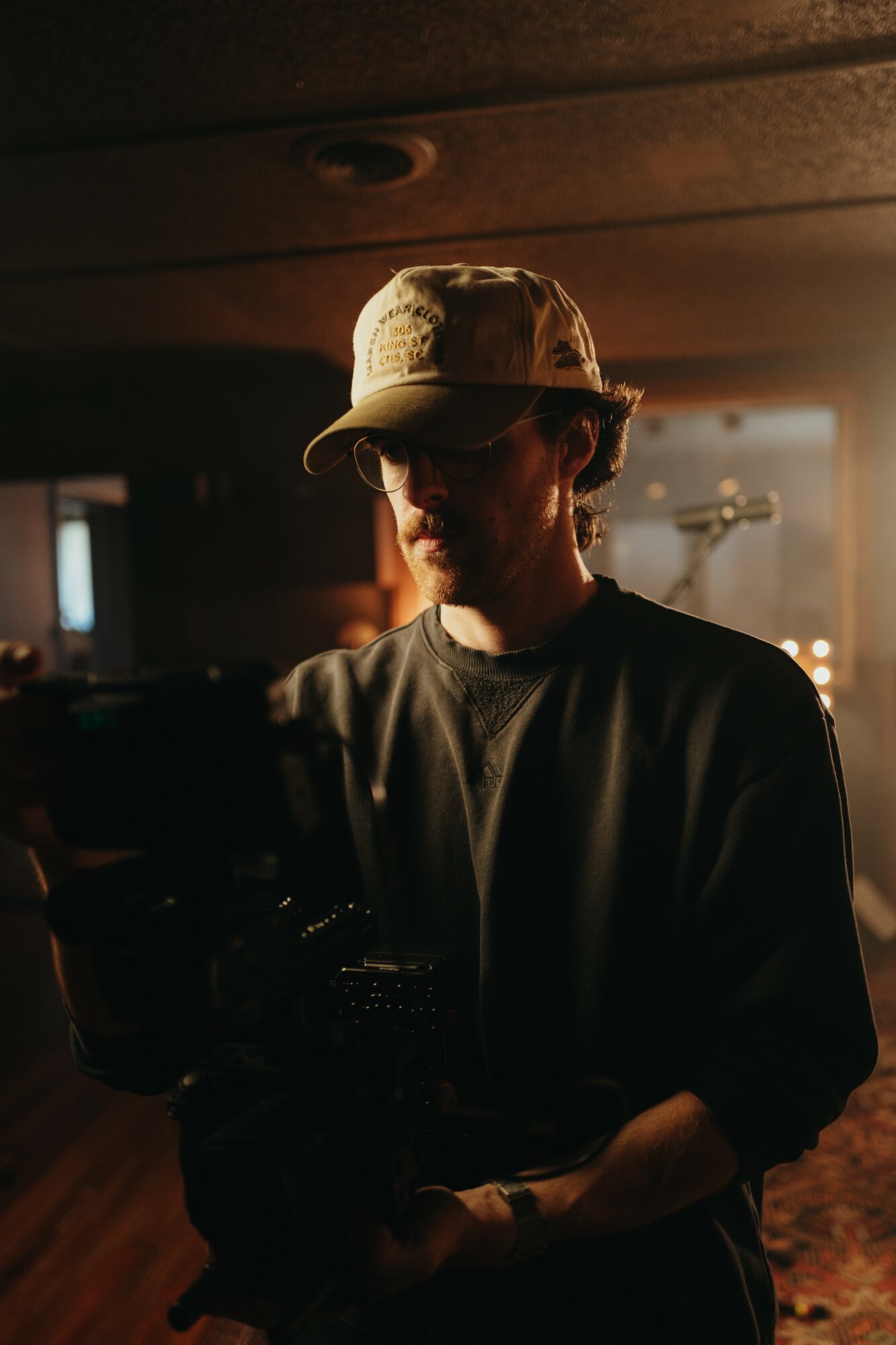 Person wearing a cap and dark clothing, standing in a dimly lit room with warm light behind.