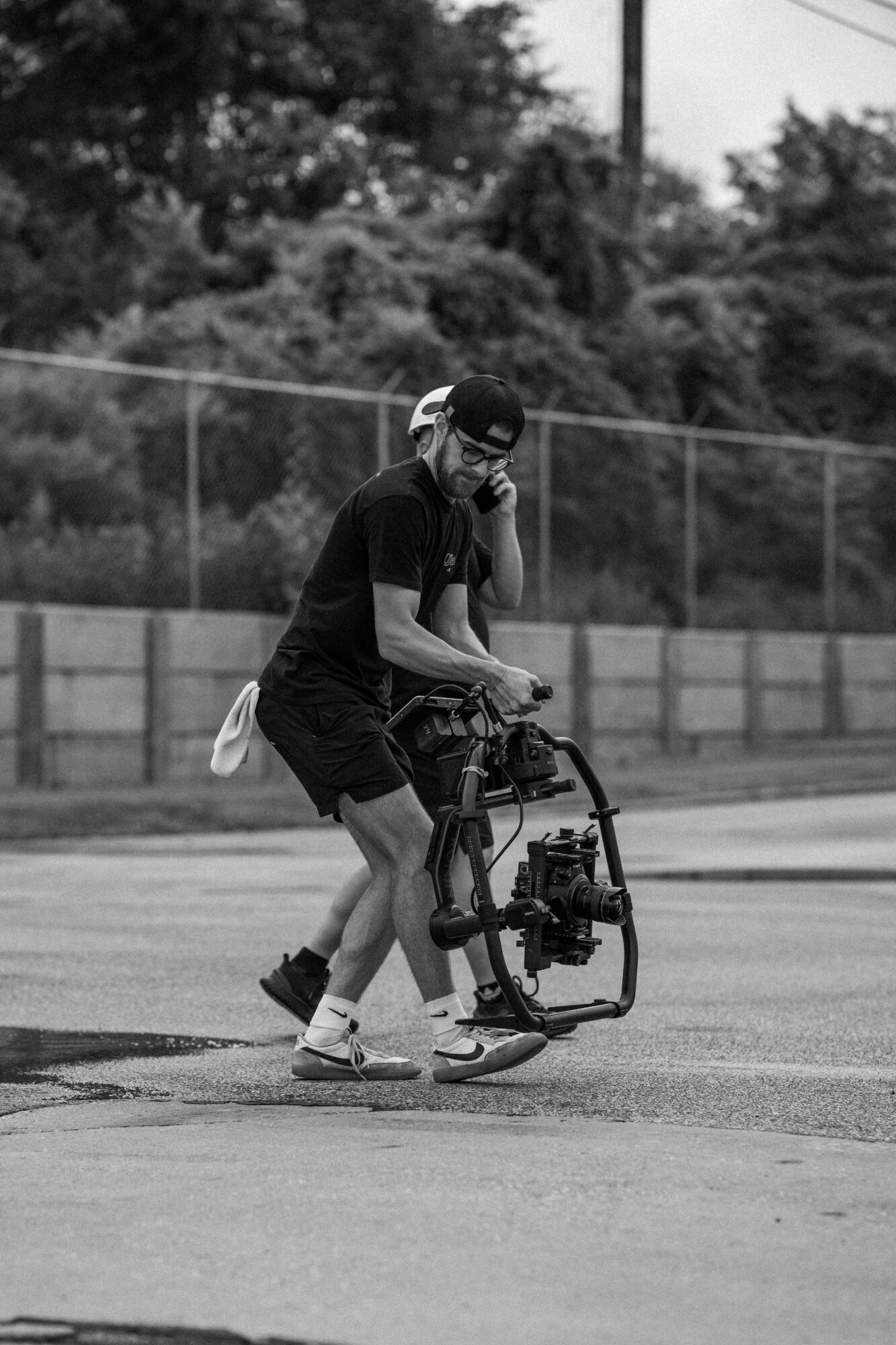Person walking on a paved surface with a camera stabilizer, wearing a helmet and casual clothing, outdoors with trees and a fence in background.