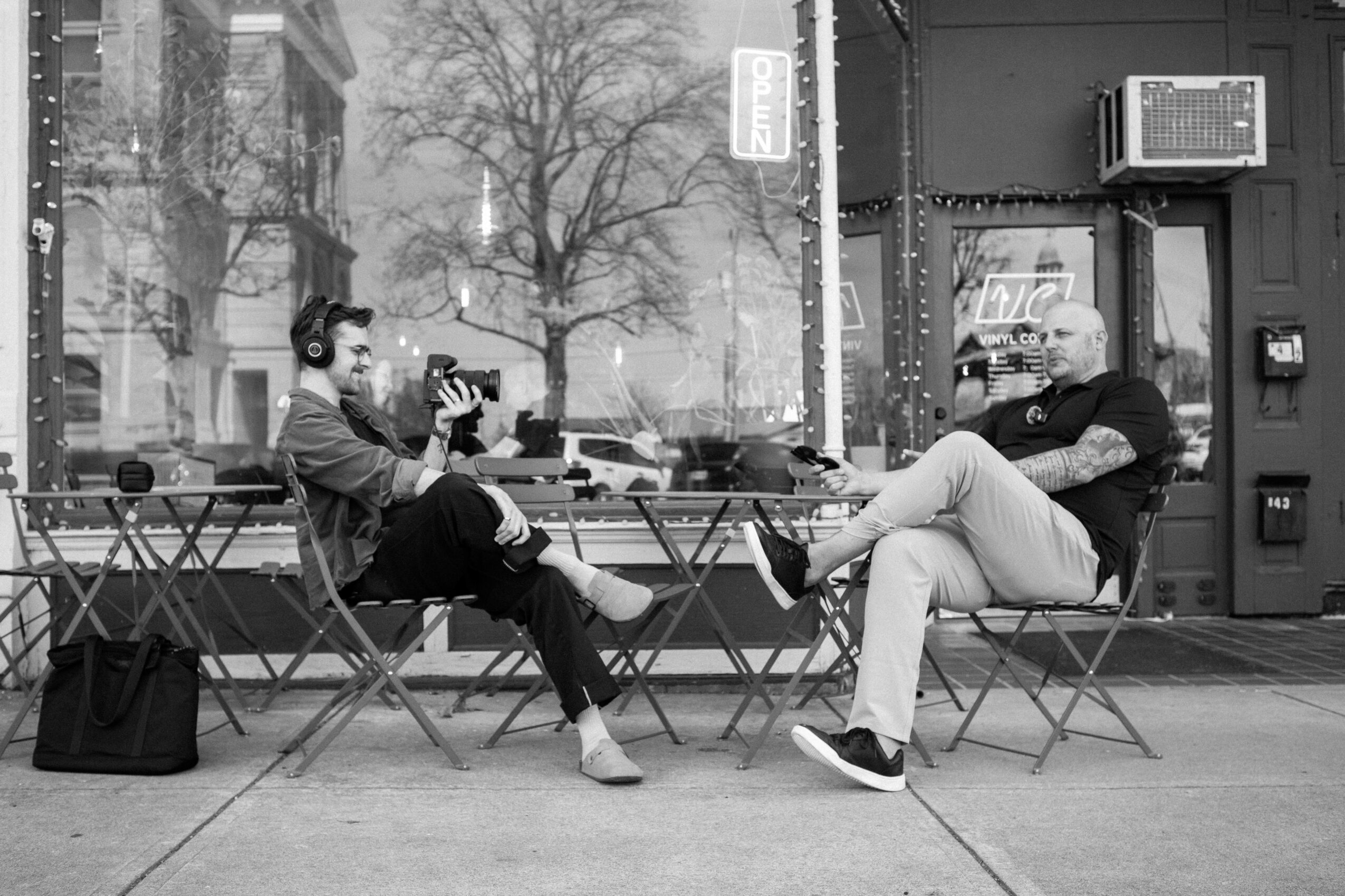 Two men sit outside a cafe, one with headphones and a camera, the other with sunglasses, on a city street.