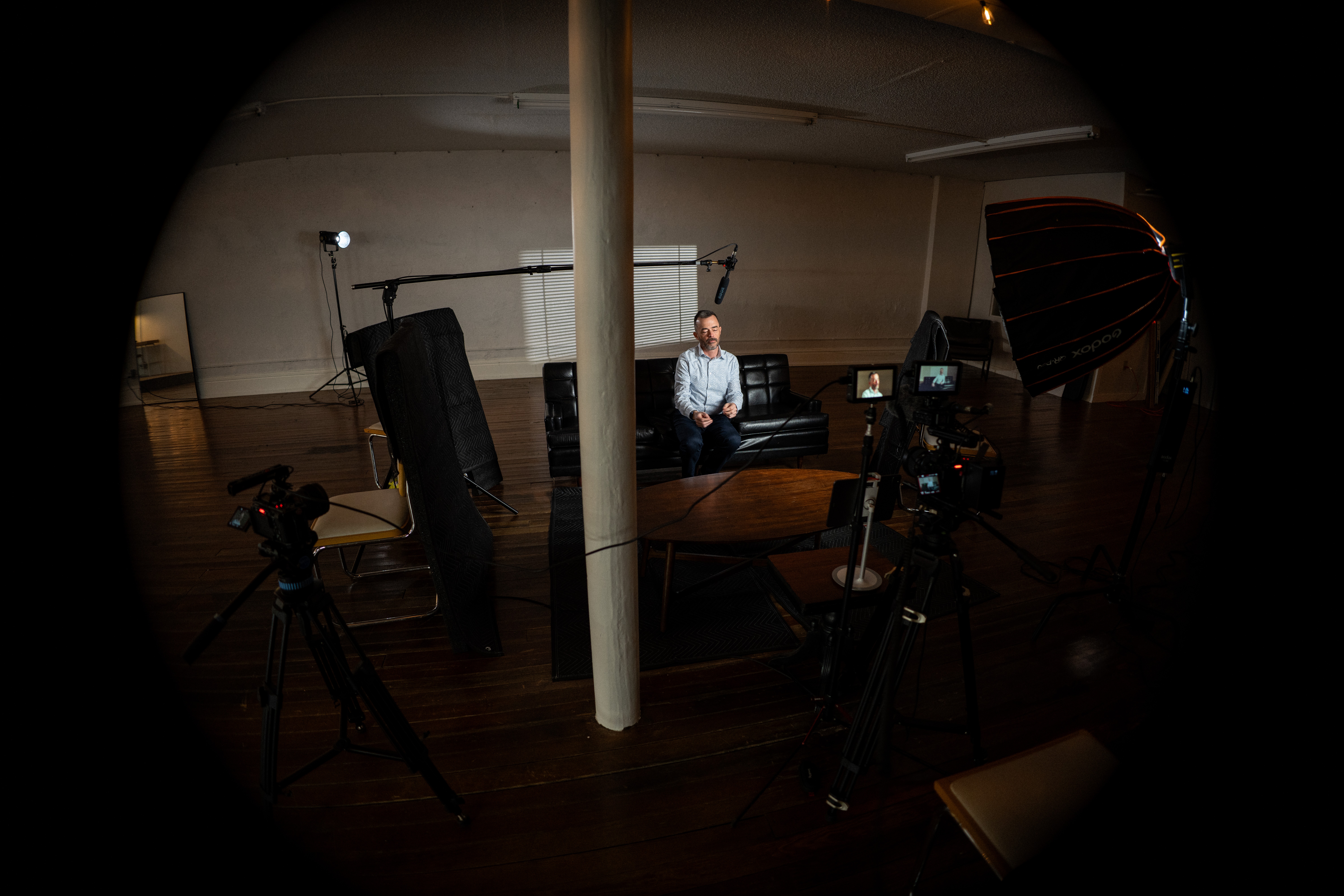 Person sitting in a studio with cameras and lighting equipment, behind a white pole, in a dimly lit room.