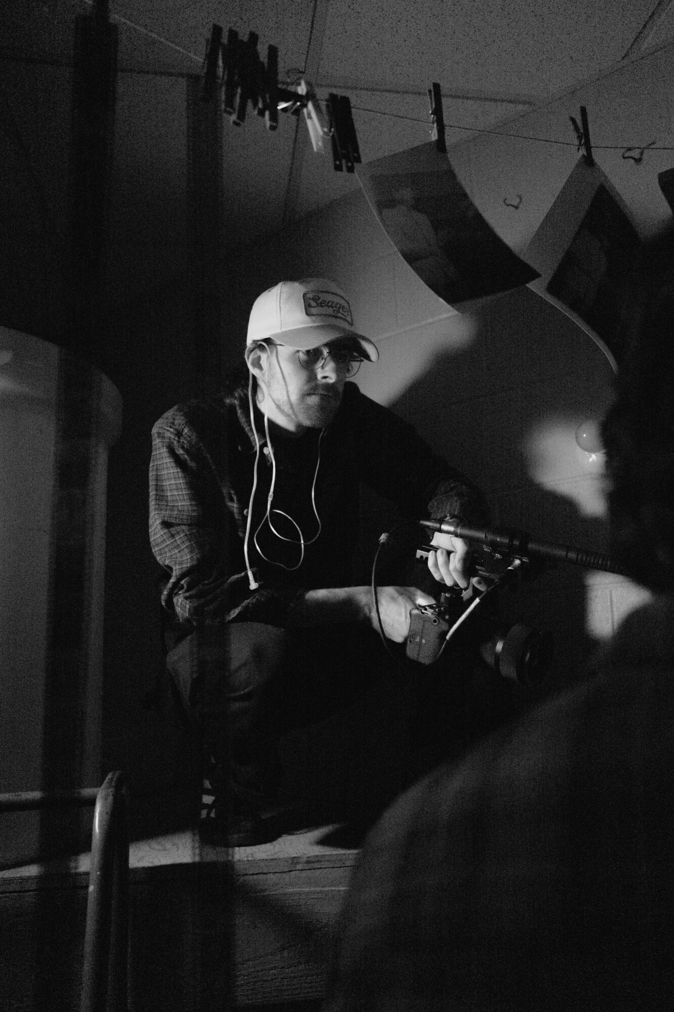 Person wearing a cap and headphones, sitting indoors with hanging photos and clothespins in the background.