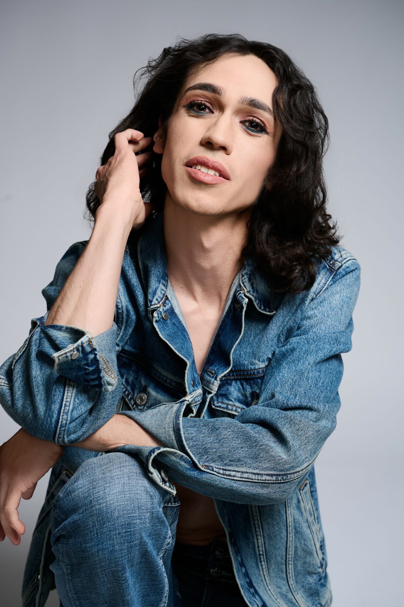 Woman with dark curly hair wearing a denim jacket, looking at the camera with a neutral expression, gray background.