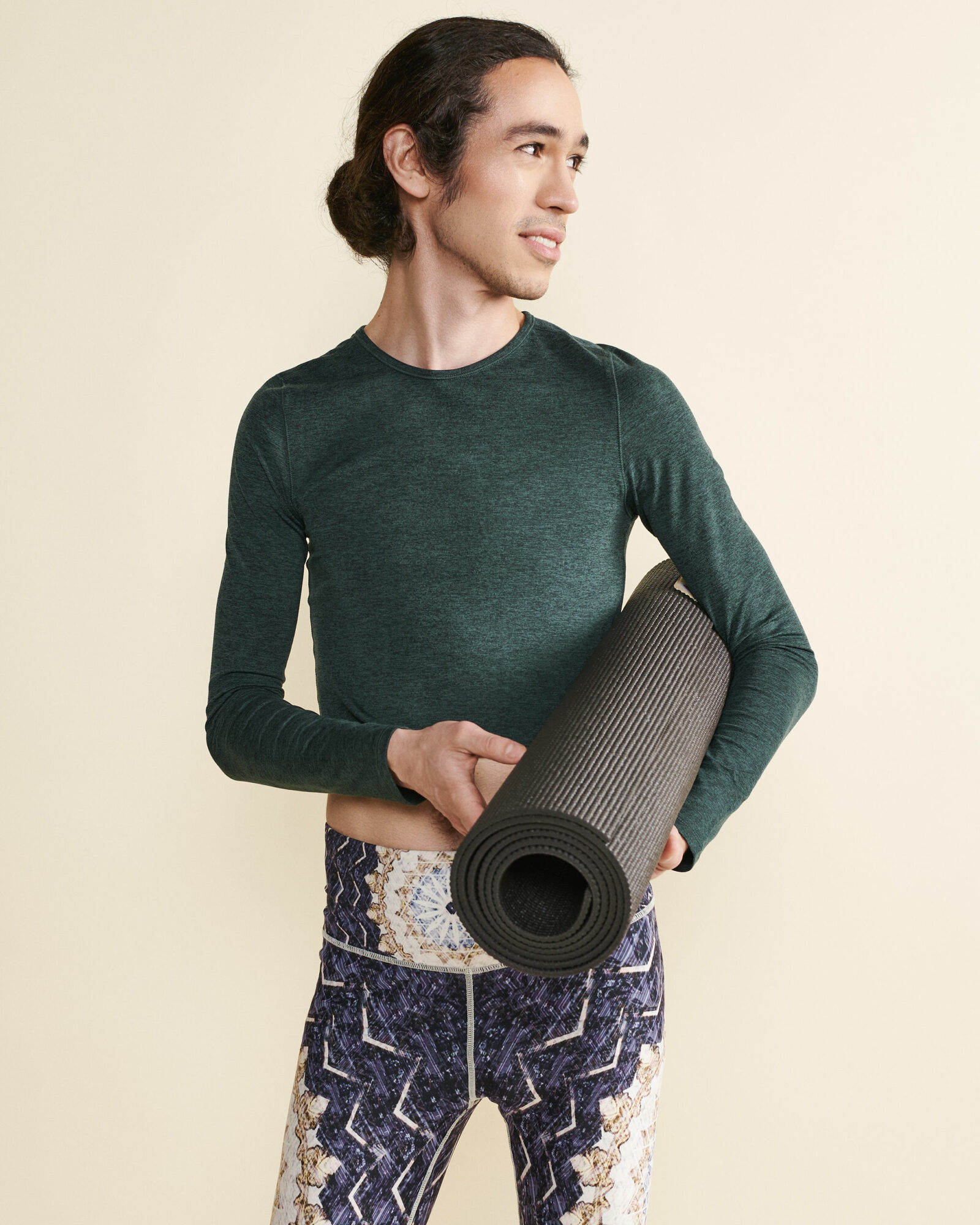 Woman holding a yoga mat, wearing a dark green long-sleeve top and patterned pants, standing against a light background.