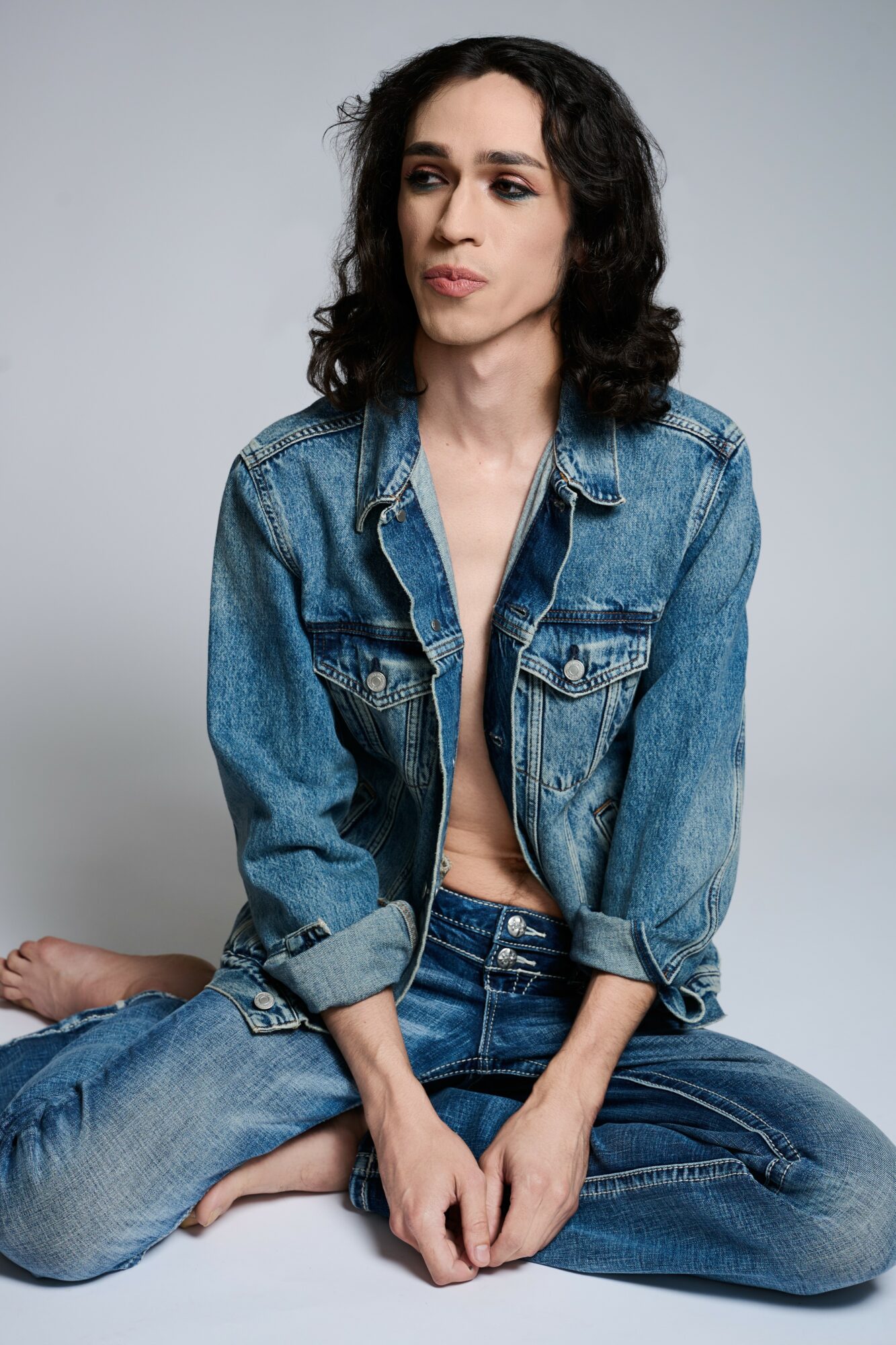 Woman with dark curly hair wearing a denim jacket and jeans, sitting cross-legged against a plain gray background.