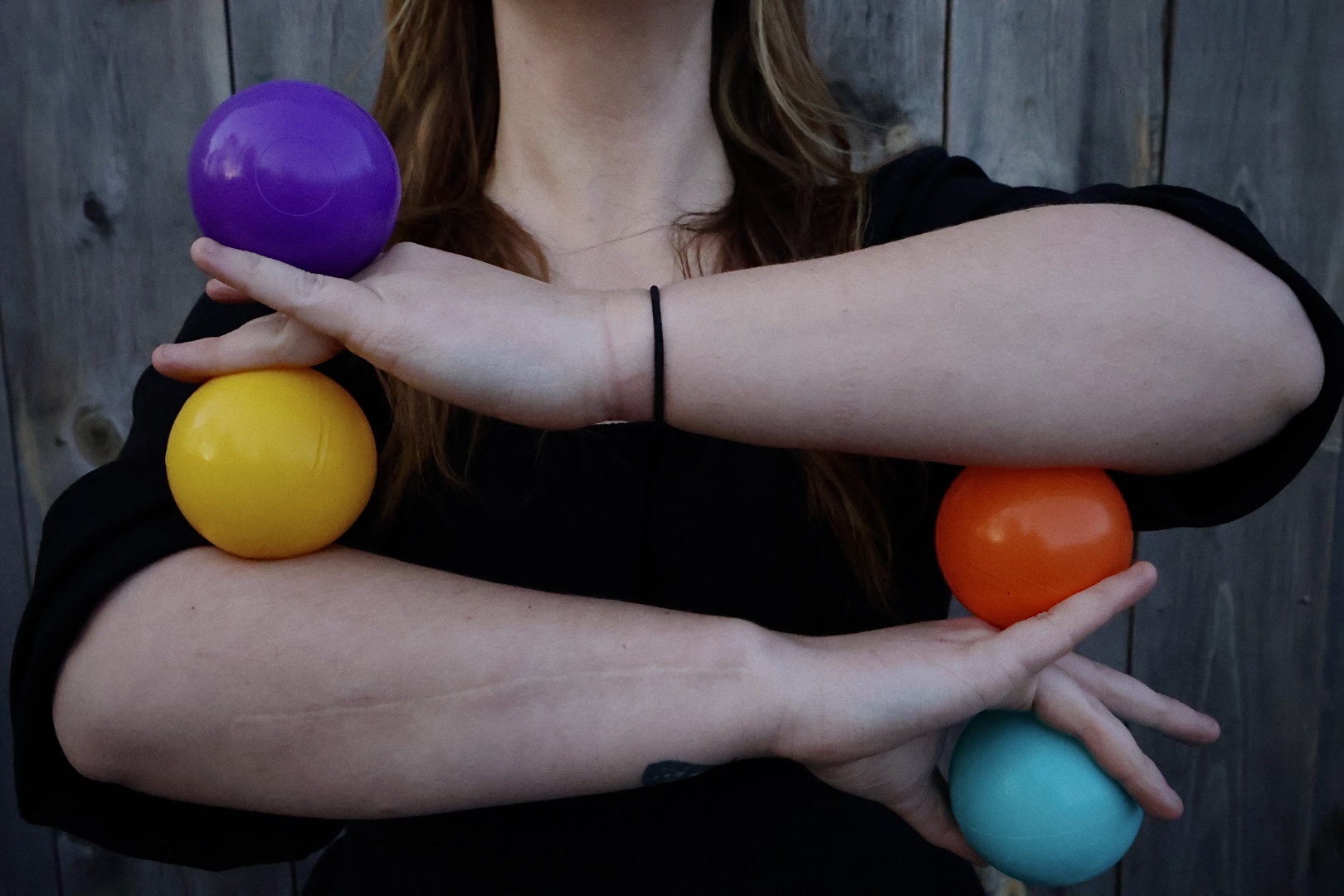 Person holding colorful balls with arms crossed, background of wooden fence, black shirt, long hair.