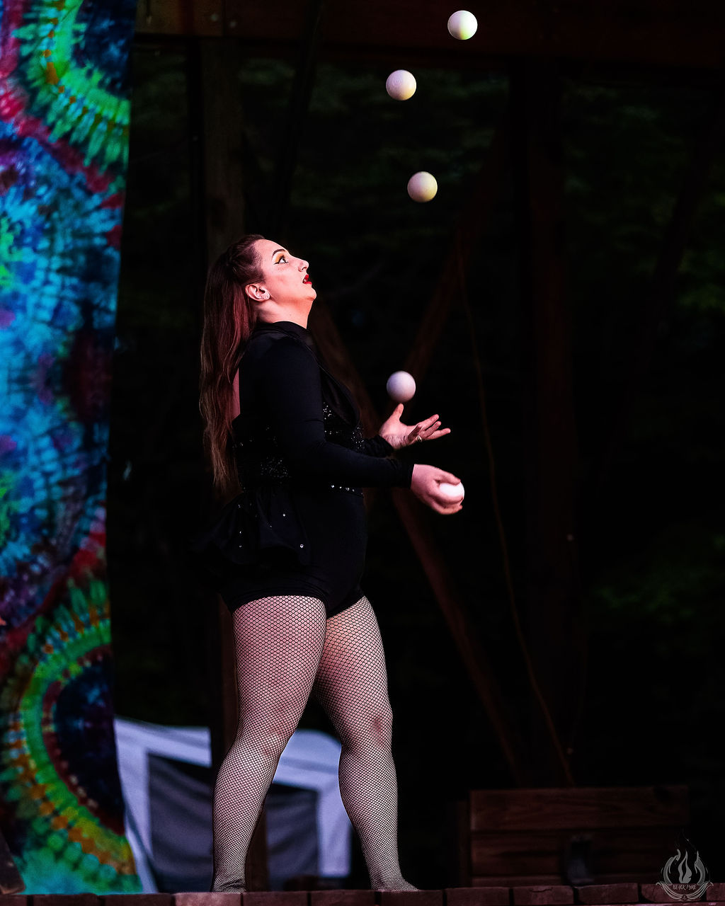 A woman juggles five balls on stage, wearing a black outfit and fishnet stockings, with a colorful backdrop behind her.