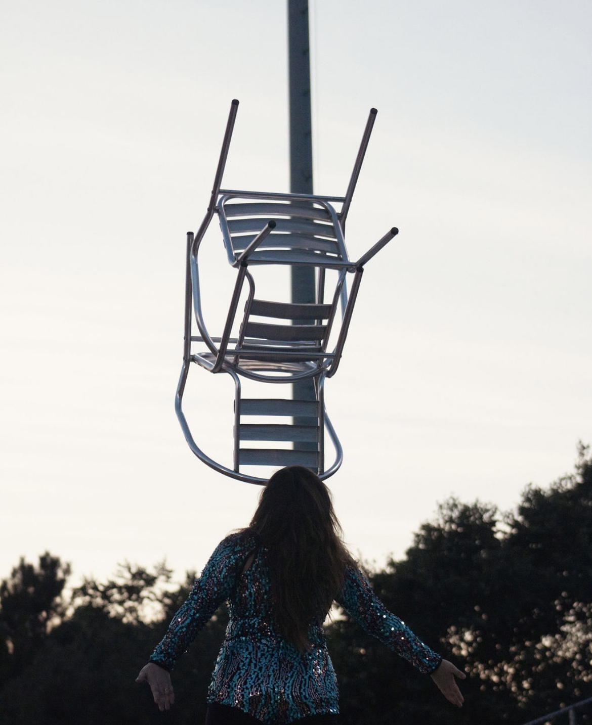 A person with long hair stands outdoors near a ladder and a pole, with trees in the background.