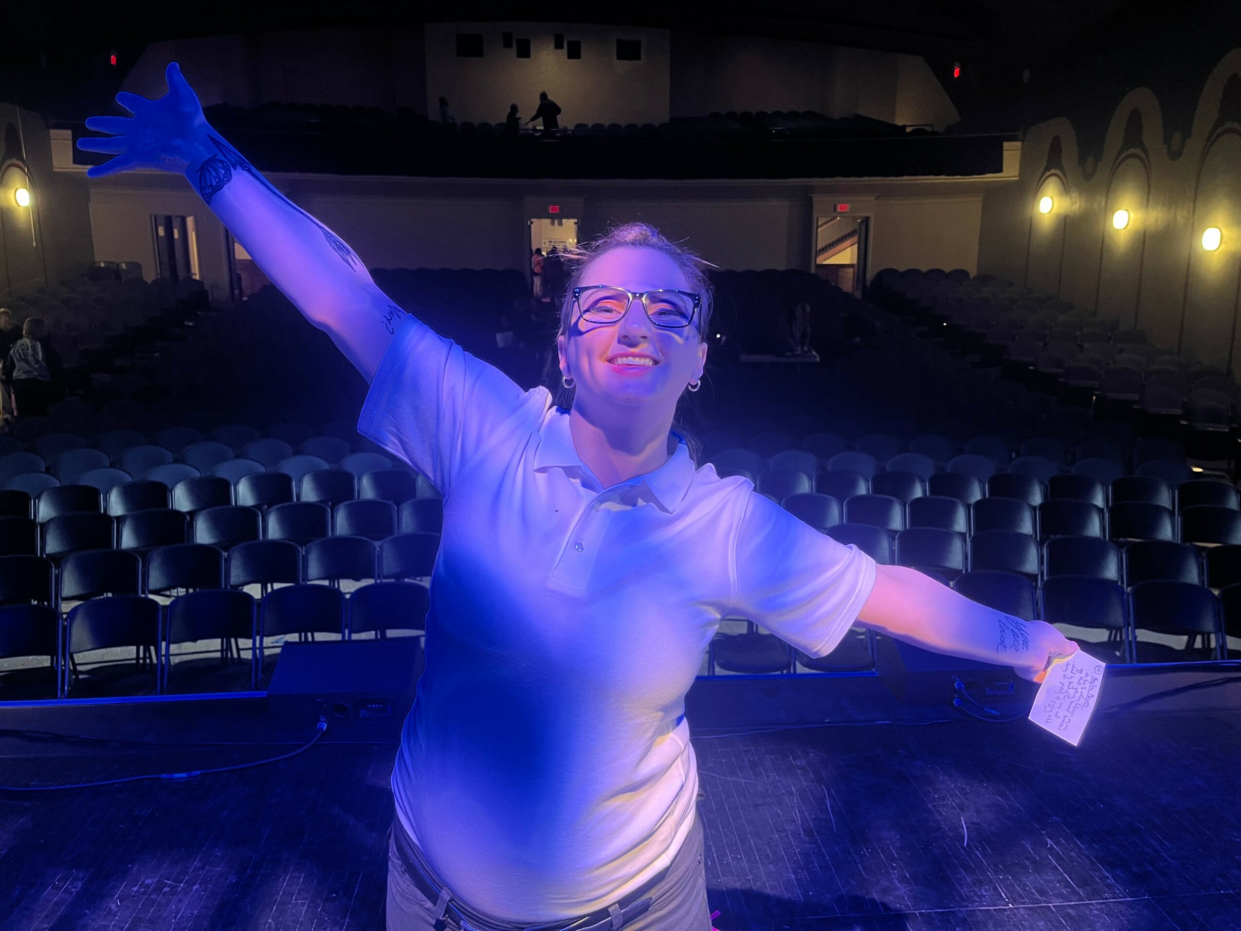 Person on stage with arms outstretched, smiling, in a theater with empty seats, stage lighting, and a dark background.