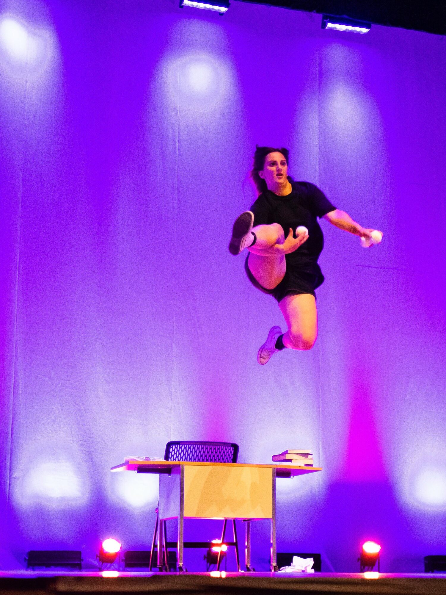 Performer mid-air on stage with purple lighting, holding a pink object, above a table with chairs.
