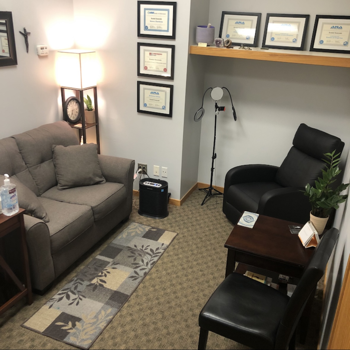 Office corner with a gray sofa, black chair, framed certificates on wall, floor lamp, and a small table with a plant.