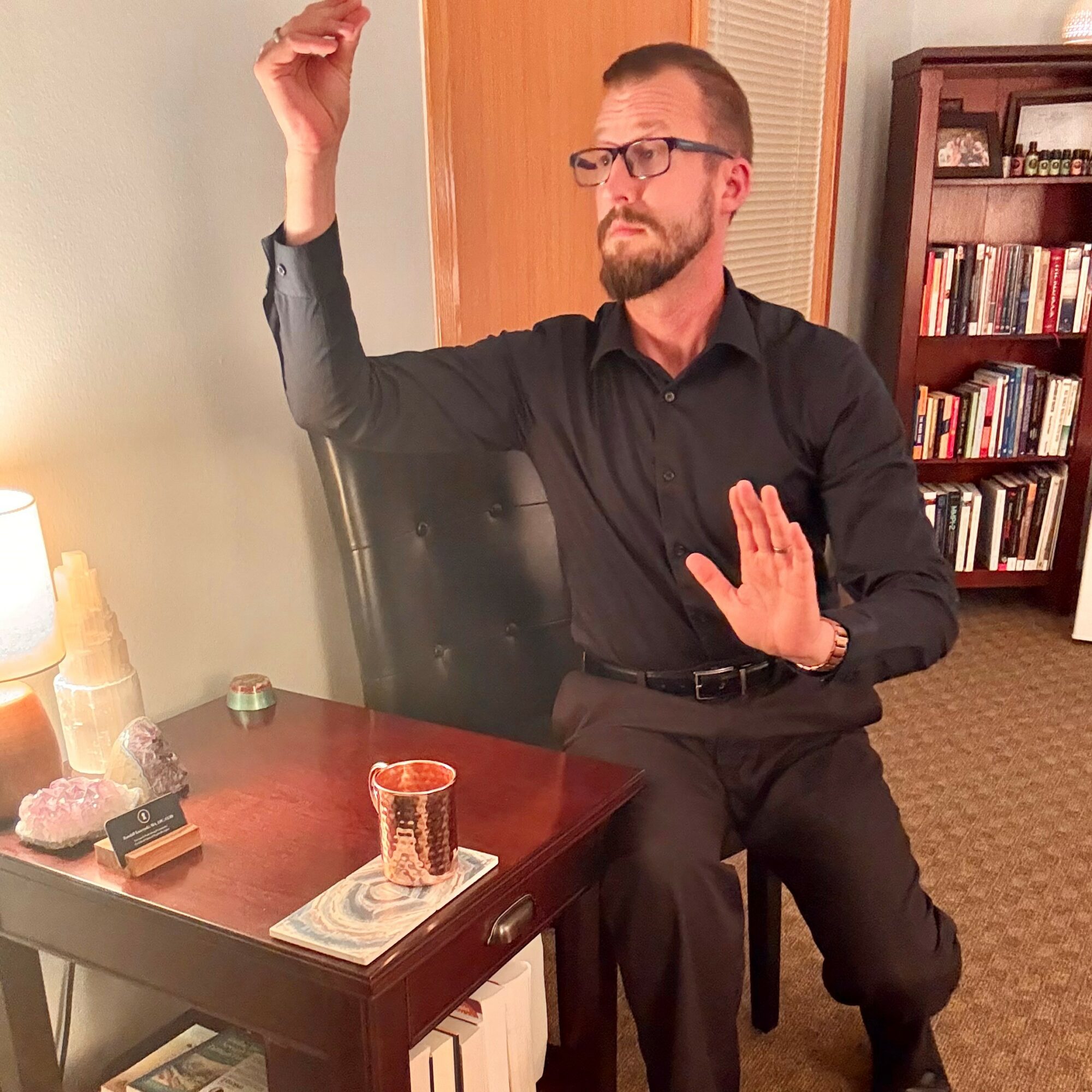 Man with glasses and beard sitting at a table, raising one hand and holding the other up, in a room with bookshelves.