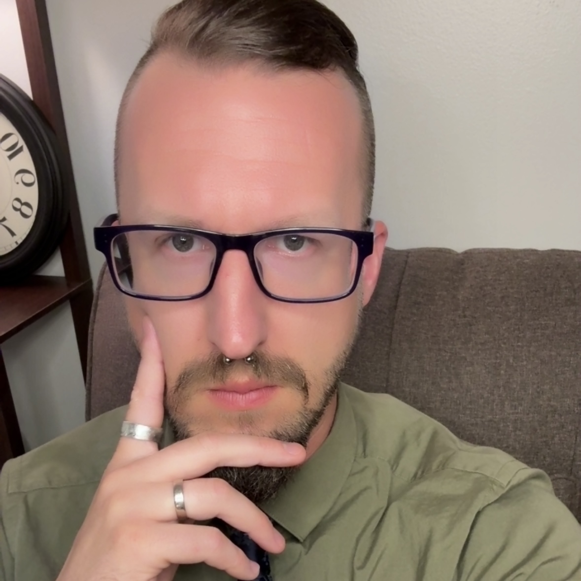 Person with glasses and a septum piercing, resting their chin on their hand, sitting on a couch.